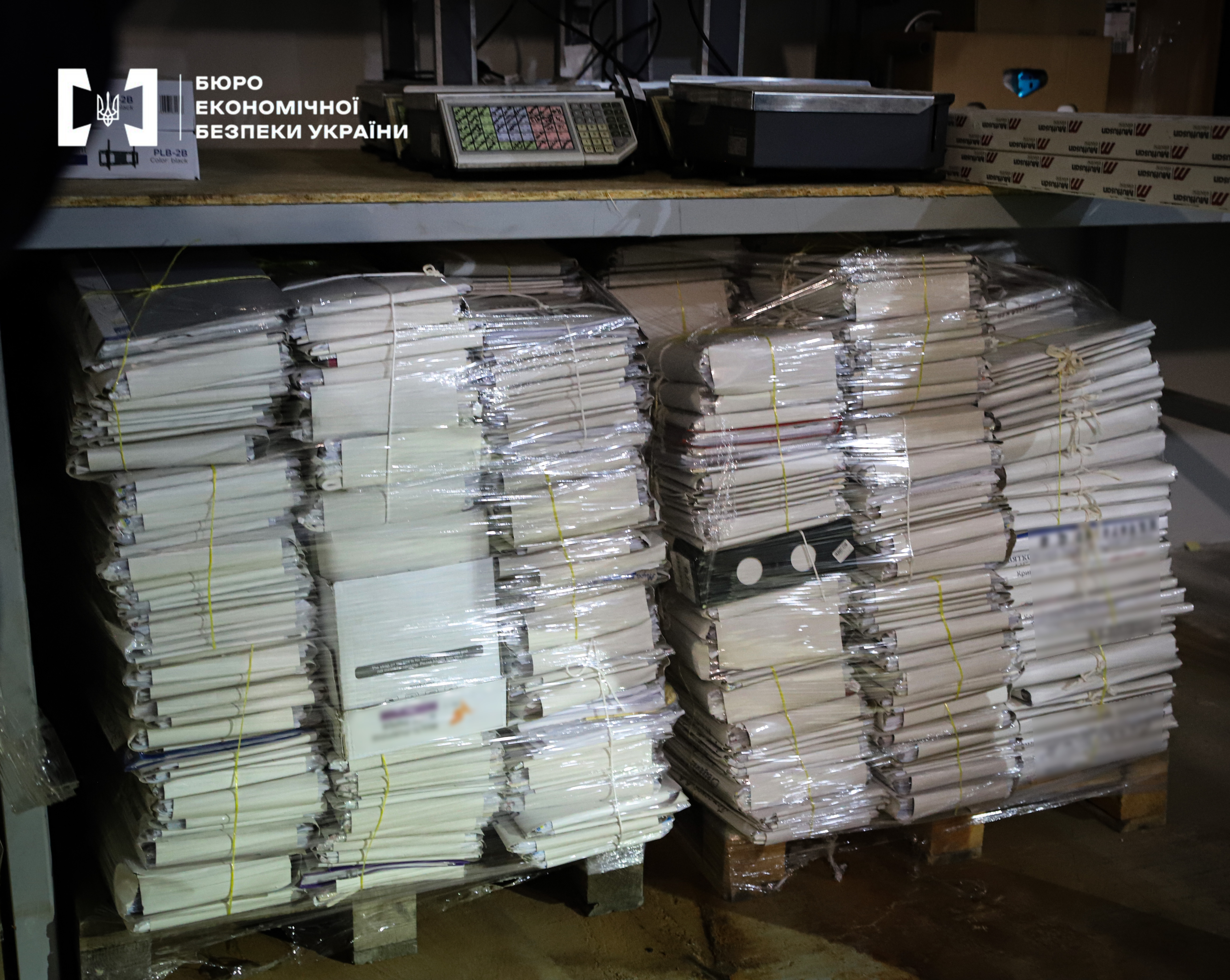 Two large pallets, fully loaded with folders and papers and tightly wrapped in transparent stretch film. Electronic scales are on a shelf above them.