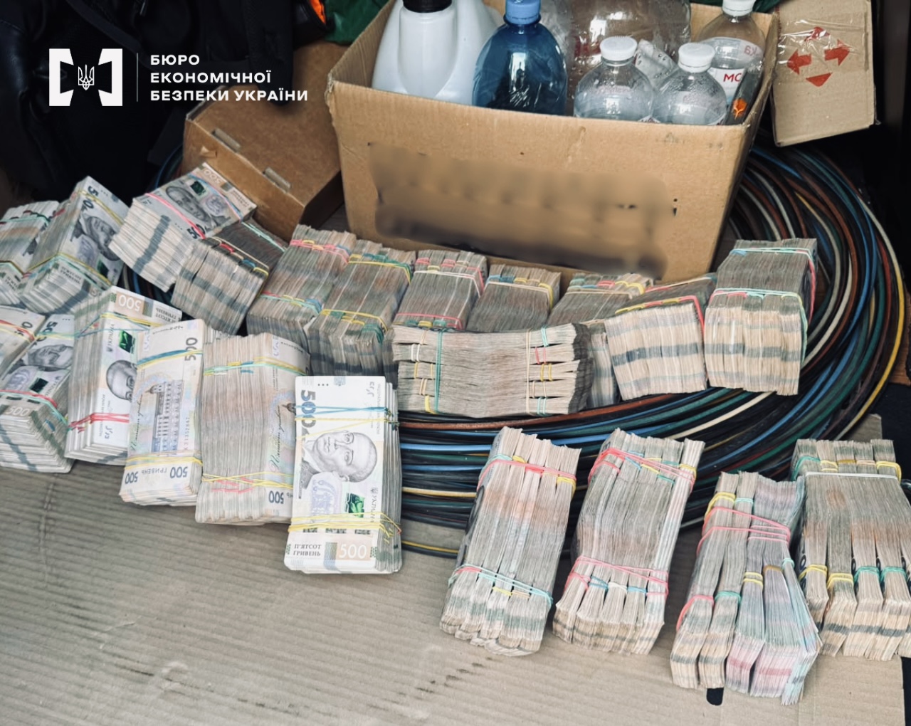 Пачки гривень (номіналом 500) розкладені на картоні поруч із бухтою кабелів. Позаду картонна коробка з пластиковими пляшками та каністрою.