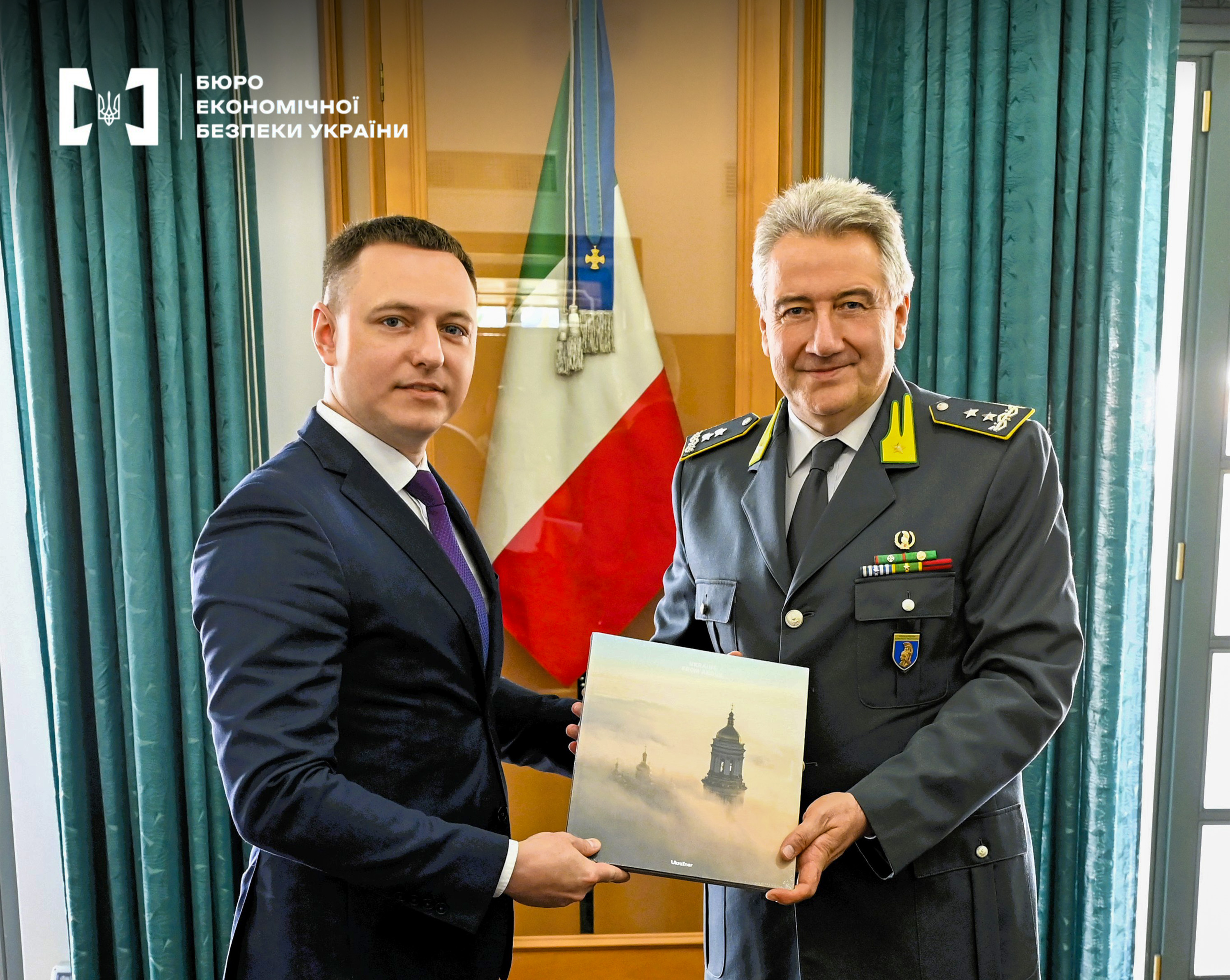 Deputy head of the ESBU Pavlo Buzdyhan and an officer of the Financial Guard of Italy in uniform hold a gift book together against the background of the Italian flag.