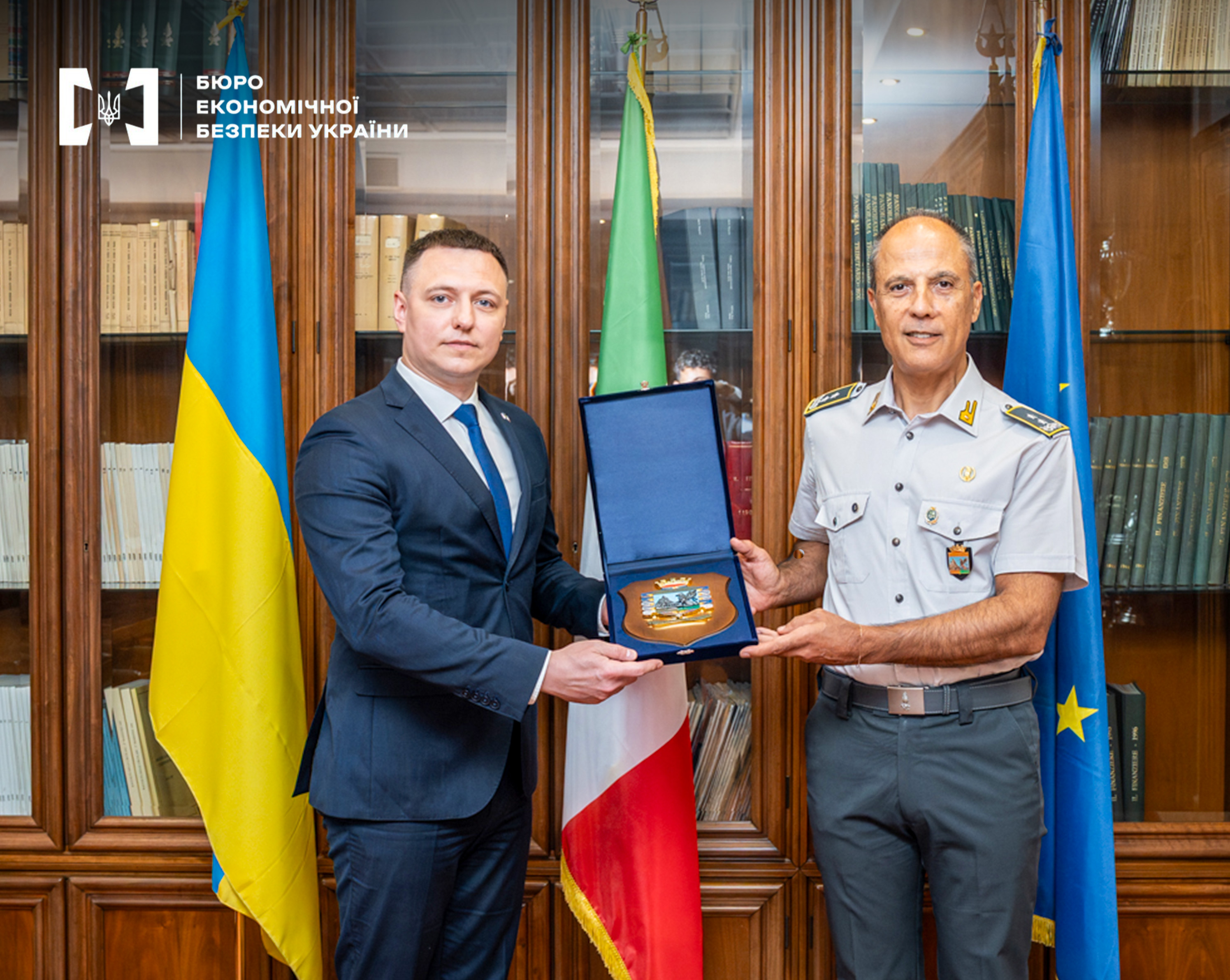 Deputy director of the ESBU and the Financial Guard of Italy with a souvenir plaque during a meeting against the background of flags and bookcases.