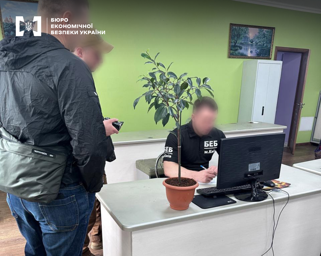 An ESBU detective sits at a computer in an office with green walls. A potted indoor plant stands in the foreground, and other men stand on the left.