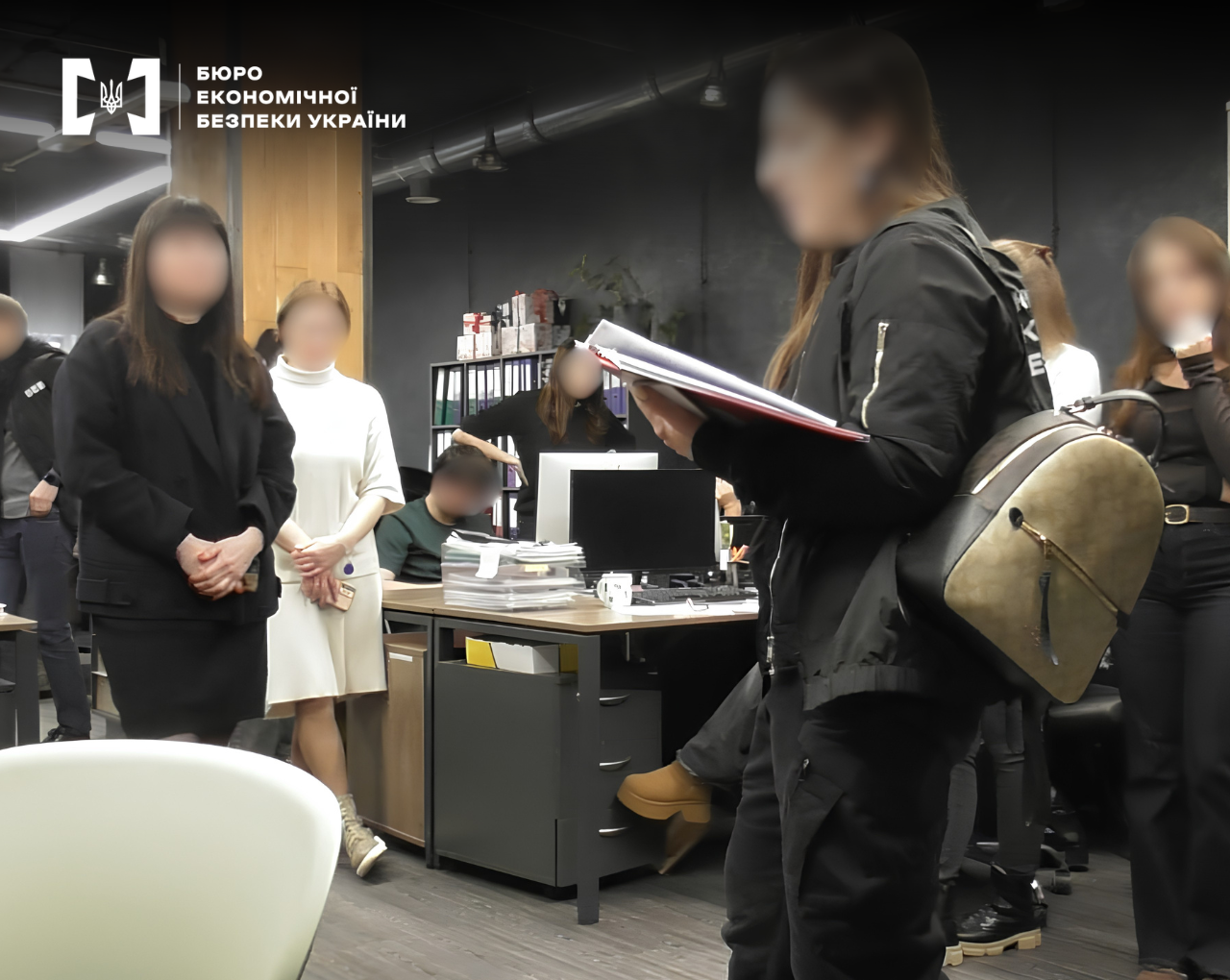 Детективи БЕБ проводять та слідчі дії в сучасному офісному приміщенні. Триває огляд документів. В кадрі багато людей, обличчя всіх заблюрено. 
