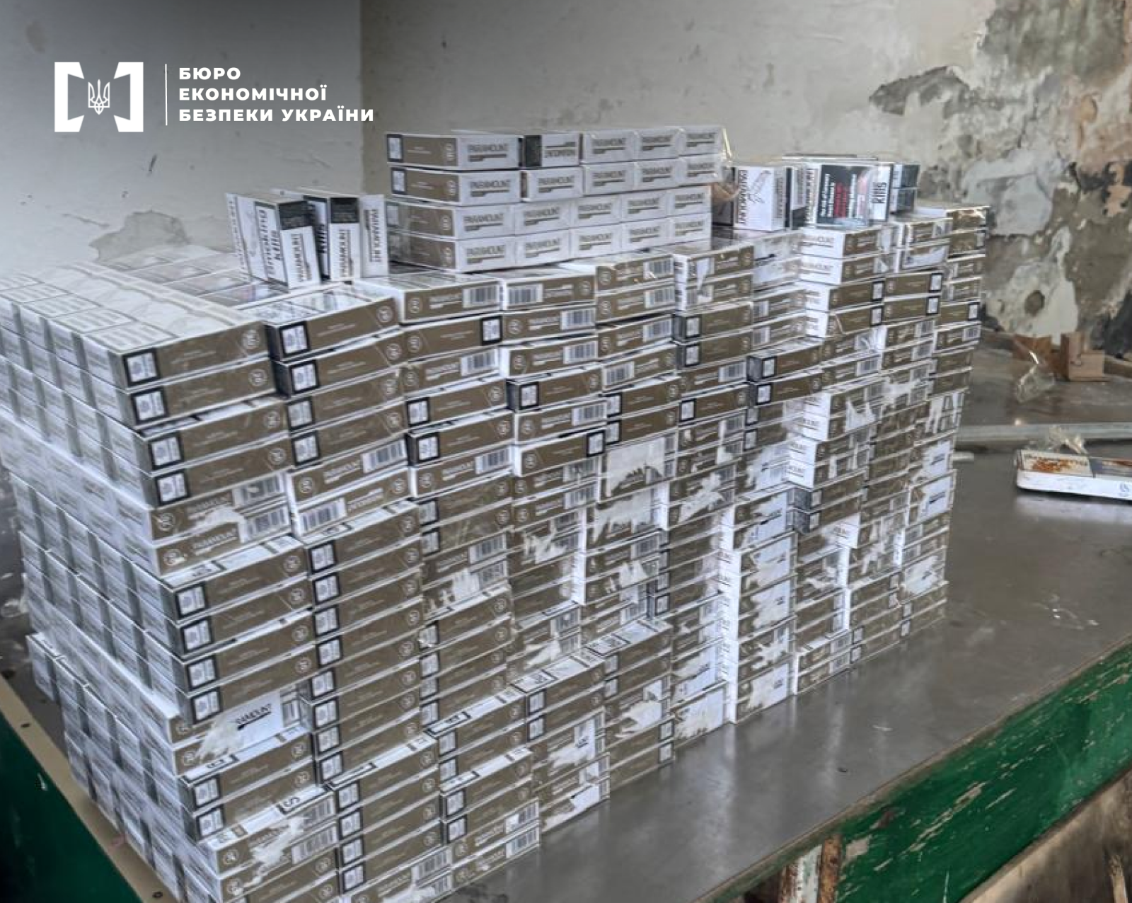 Hundreds of identical cigarette packs are stacked in high rows on a metal table in a dilapidated room.