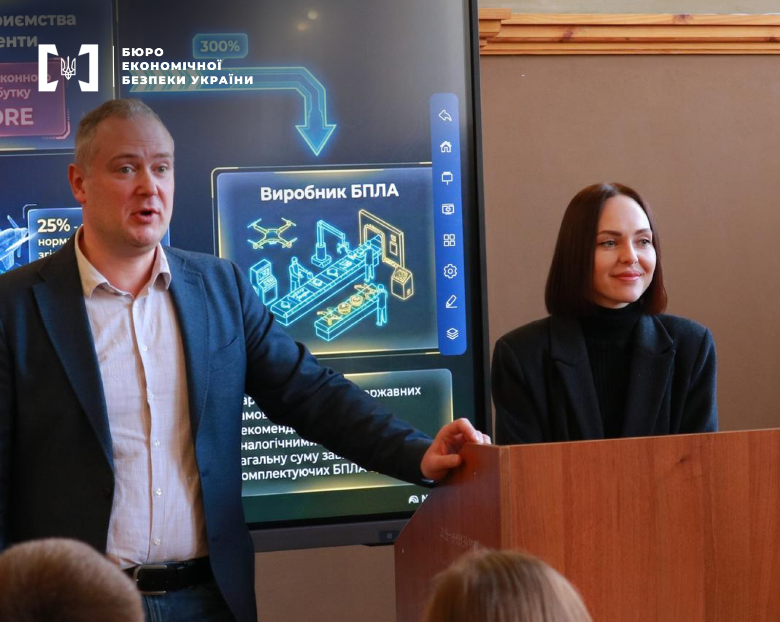 A man in a blue blazer presents a report from the ESBU. Next to him at the podium stands a woman in a black jacket. On the screen behind them is an infographic about UAV production.