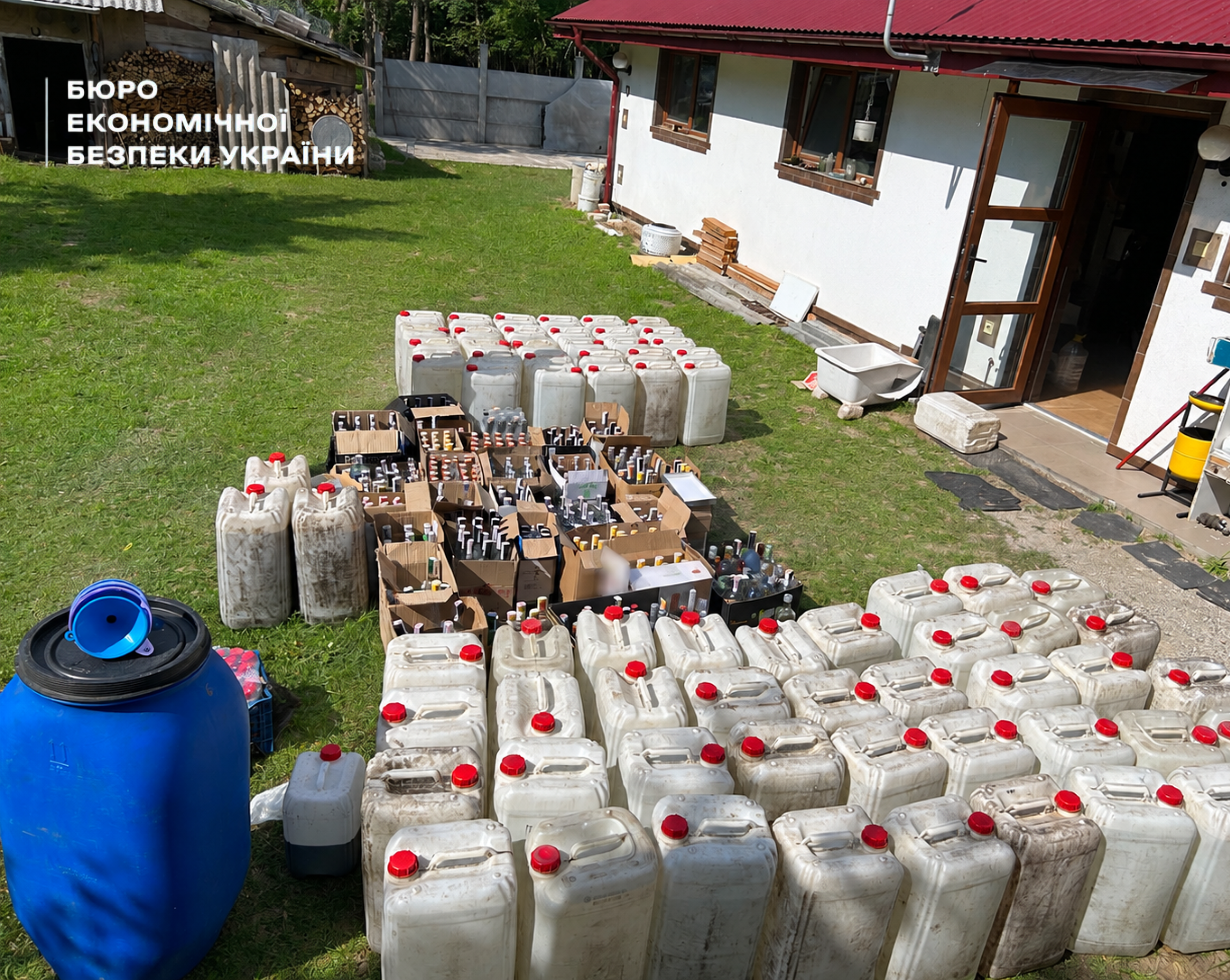 Dozens of white canisters with red caps, boxes of vodka of various brands, and a blue barrel on a lawn near a house.