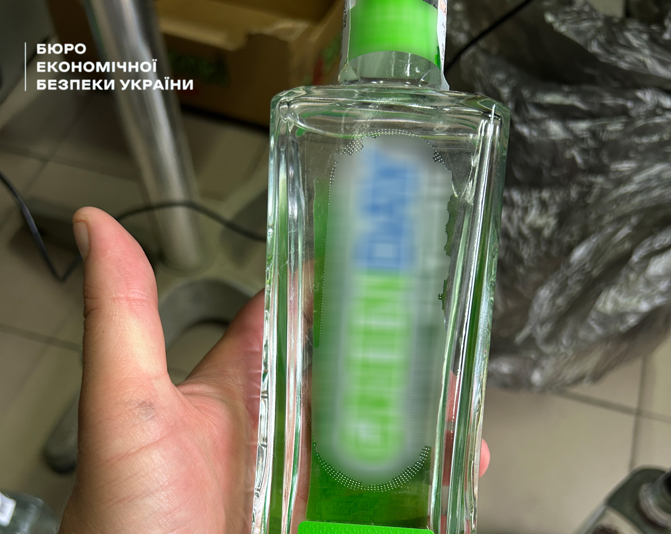 A hand holds a transparent rectangular glass bottle with a bright green label and an excise stamp on the neck. 