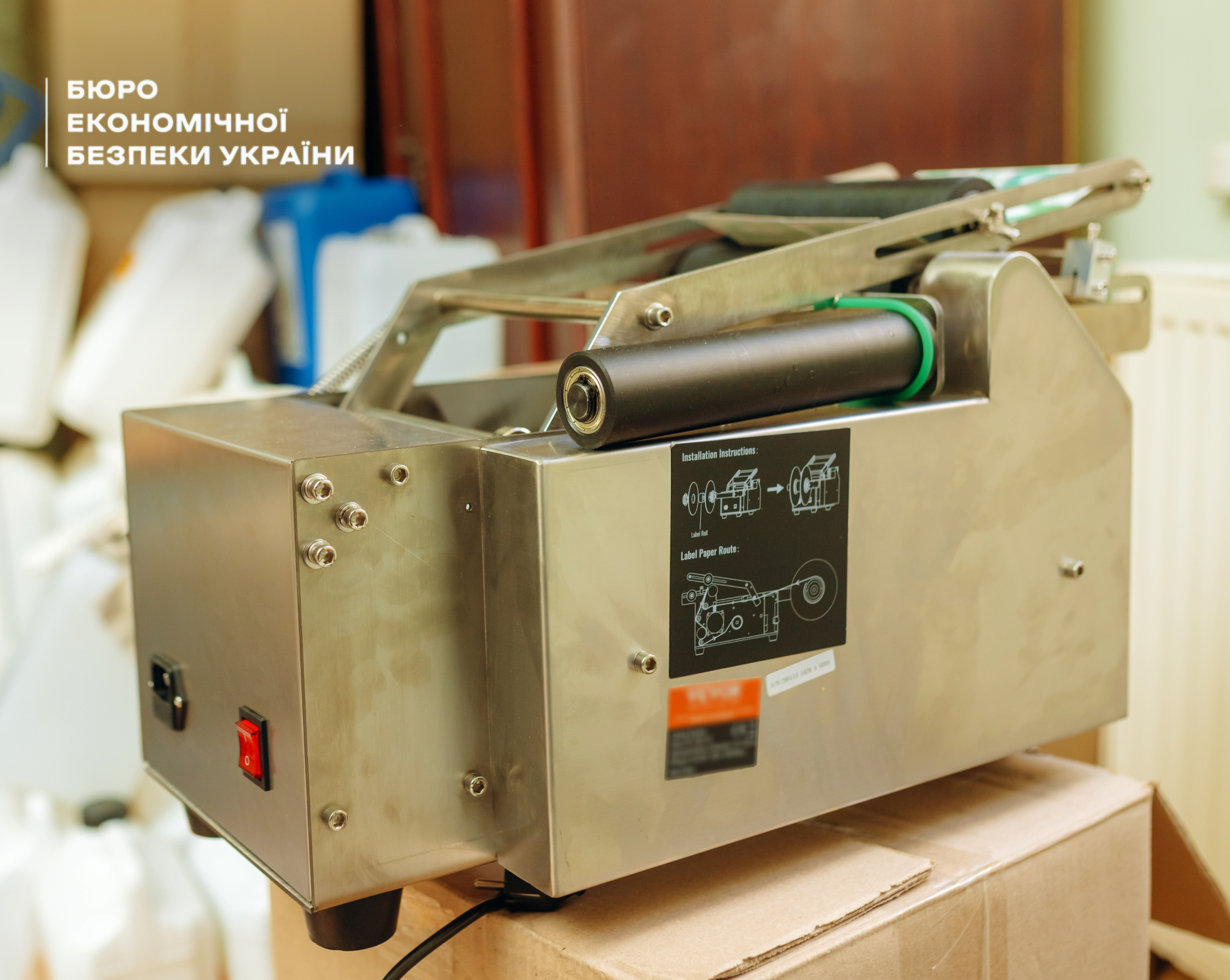 A metal semi-automatic labeling machine with rollers, instructions on the housing, and a power button, resting on a cardboard box.