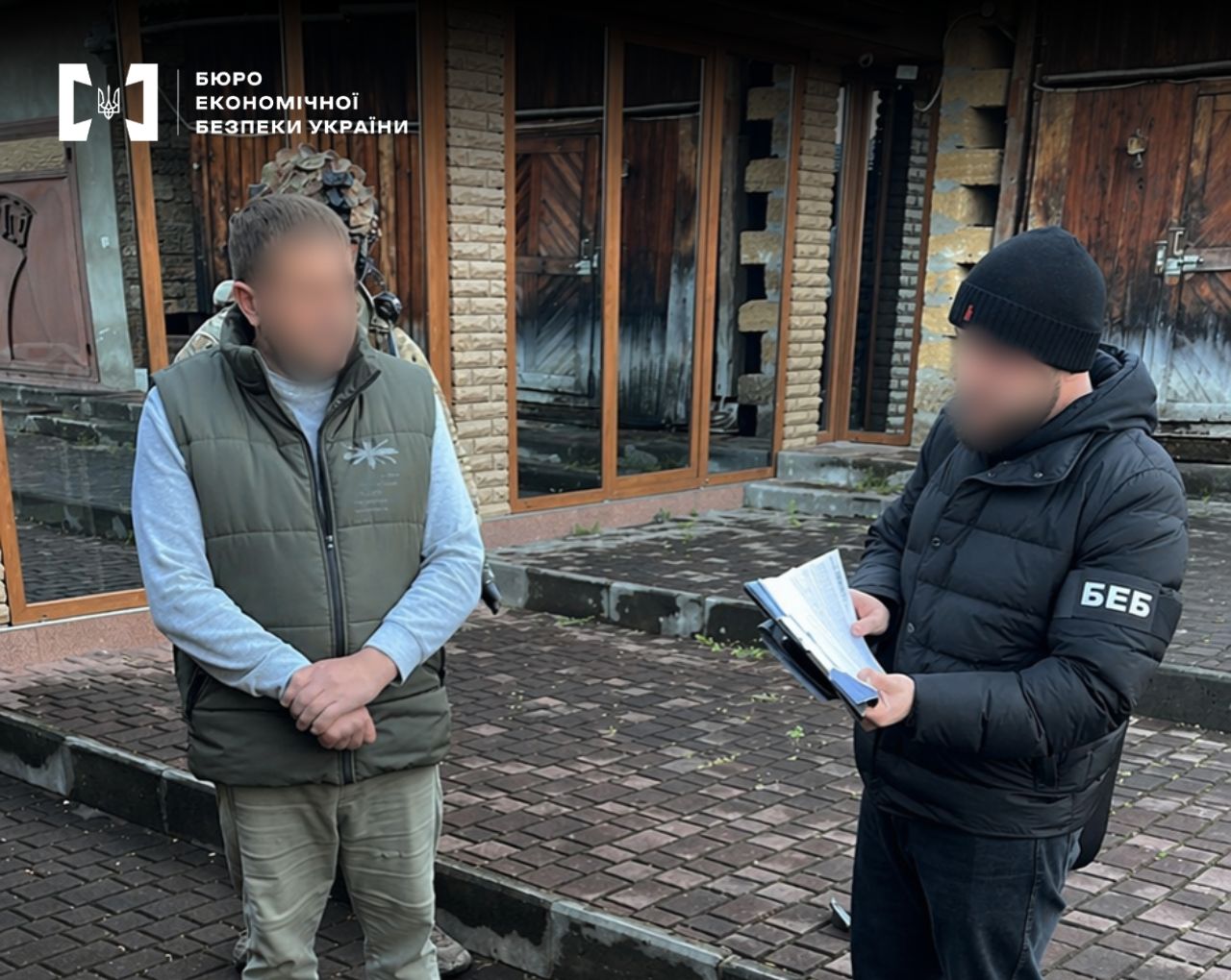 An ESBU detective is holding documents in his hands. The faces of both men are blurred. In the background, a brick building and a person in camouflage uniform are visible