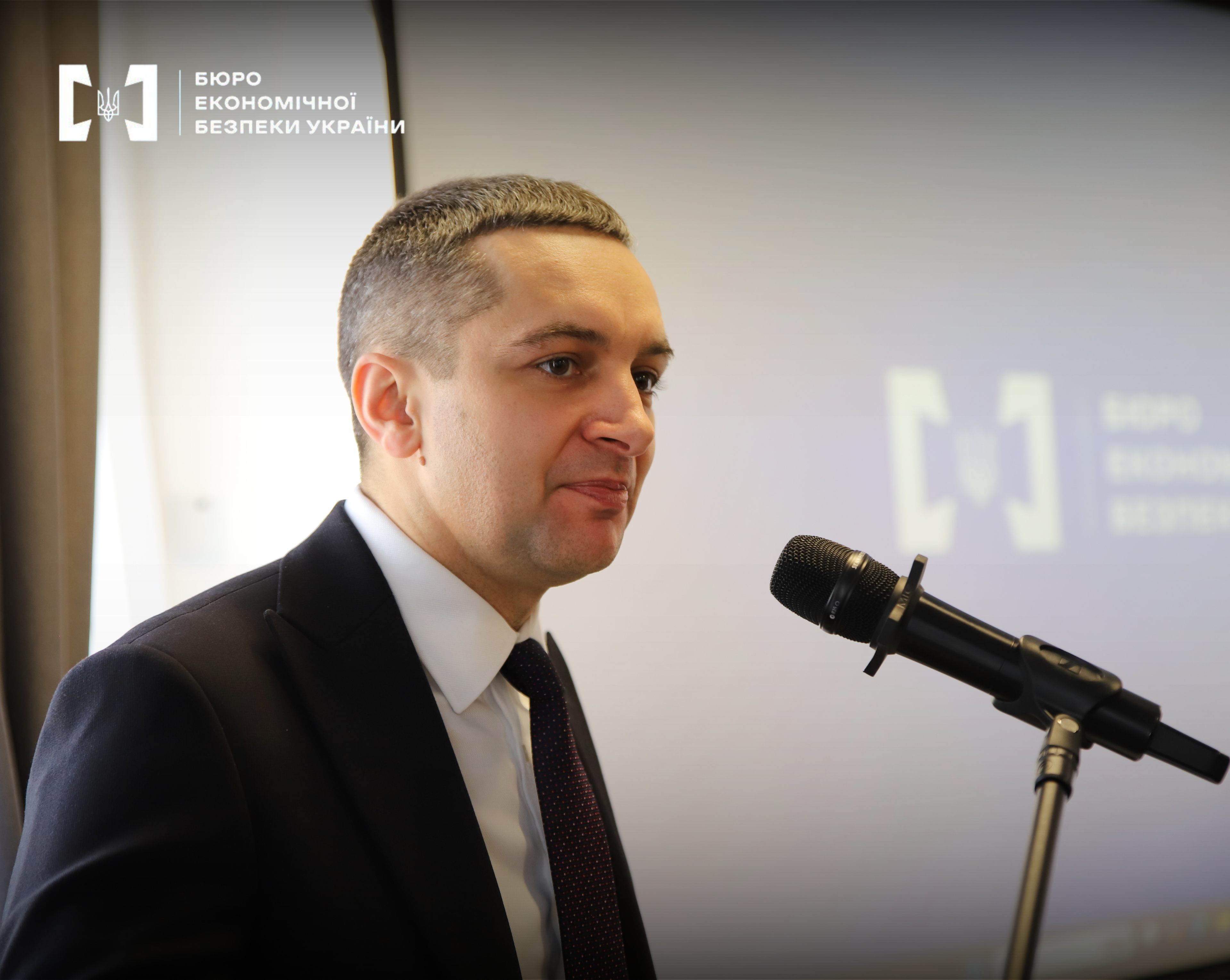 First Deputy Director of the Economic Security Bureau of Ukraine, Taras Shcherbai, stands before a microphone with the agency’s logo visible in the background