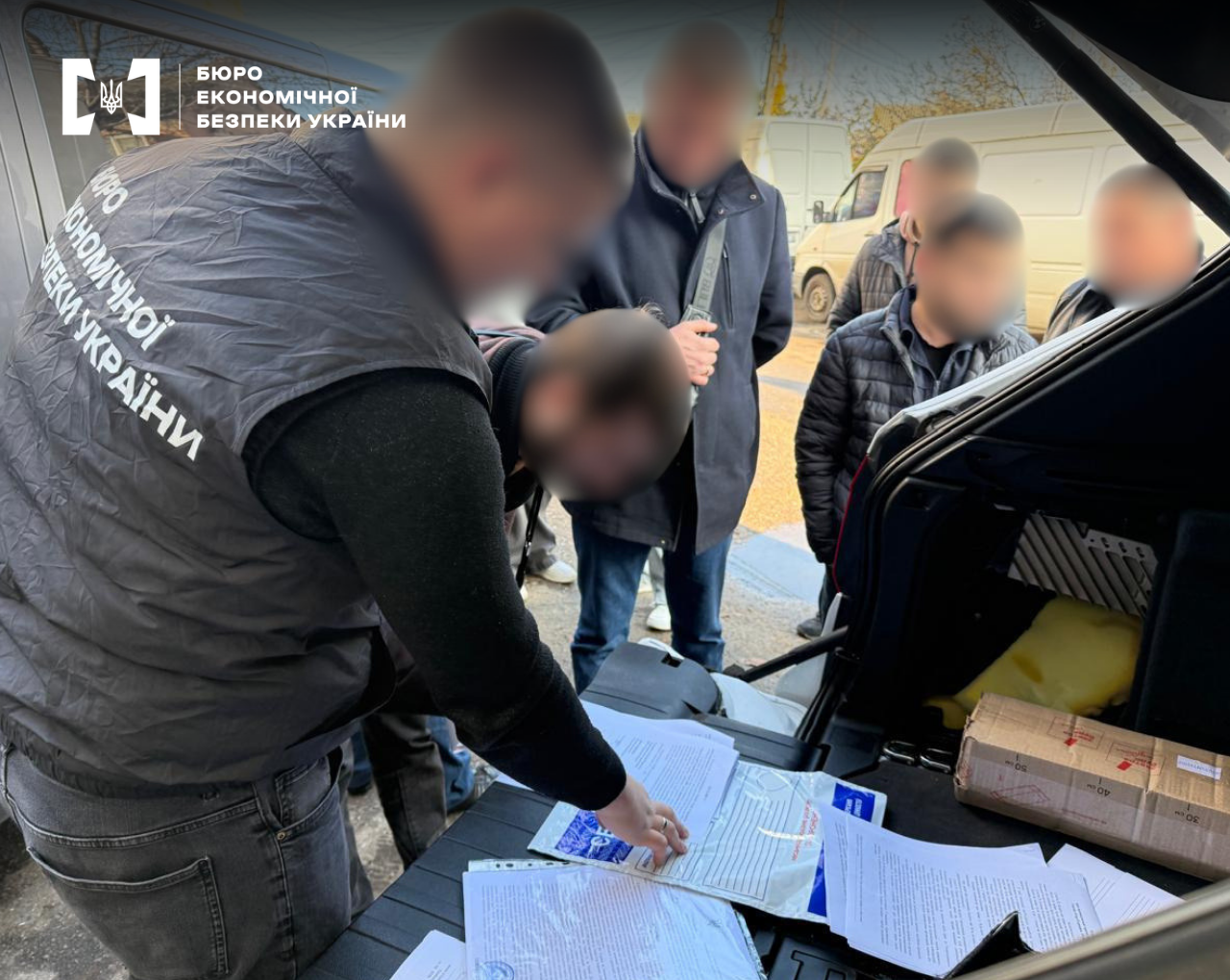 An ESBU detective inventories seized documentation during a search. Piles of papers, financial reports, and contracts are spread across the open trunk of a car. Witnesses and participants of the investigative action are present nearby