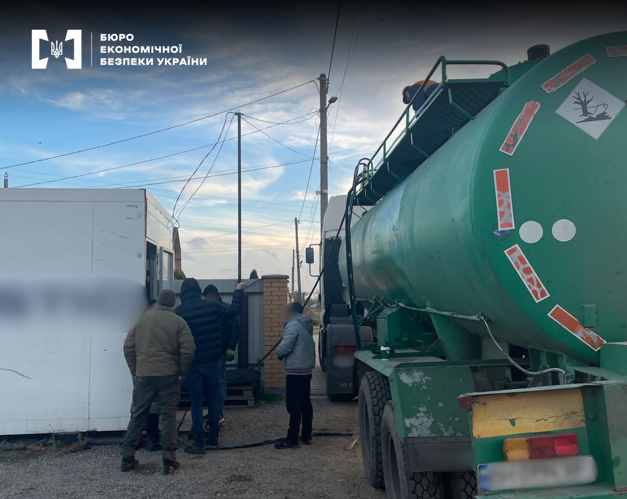 A green tank truck is parked next to a mobile white kiosk, near which several people are standing