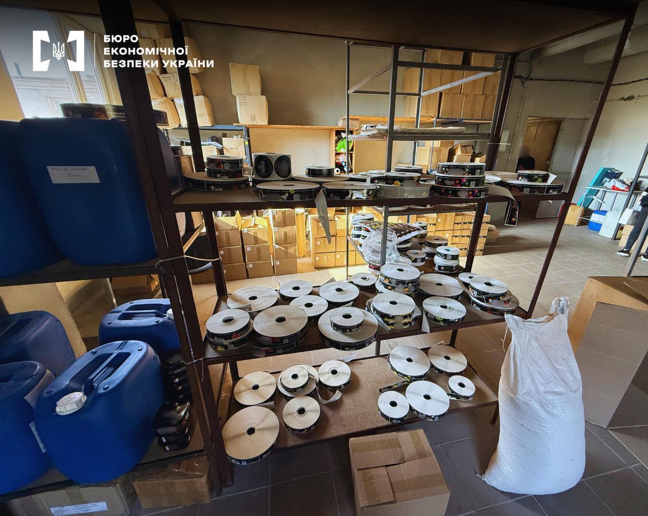 Warehouse shelving units are filled with numerous rolls of excise stamps or labels. Nearby are blue plastic canisters, a large white sack, and cardboard boxes.