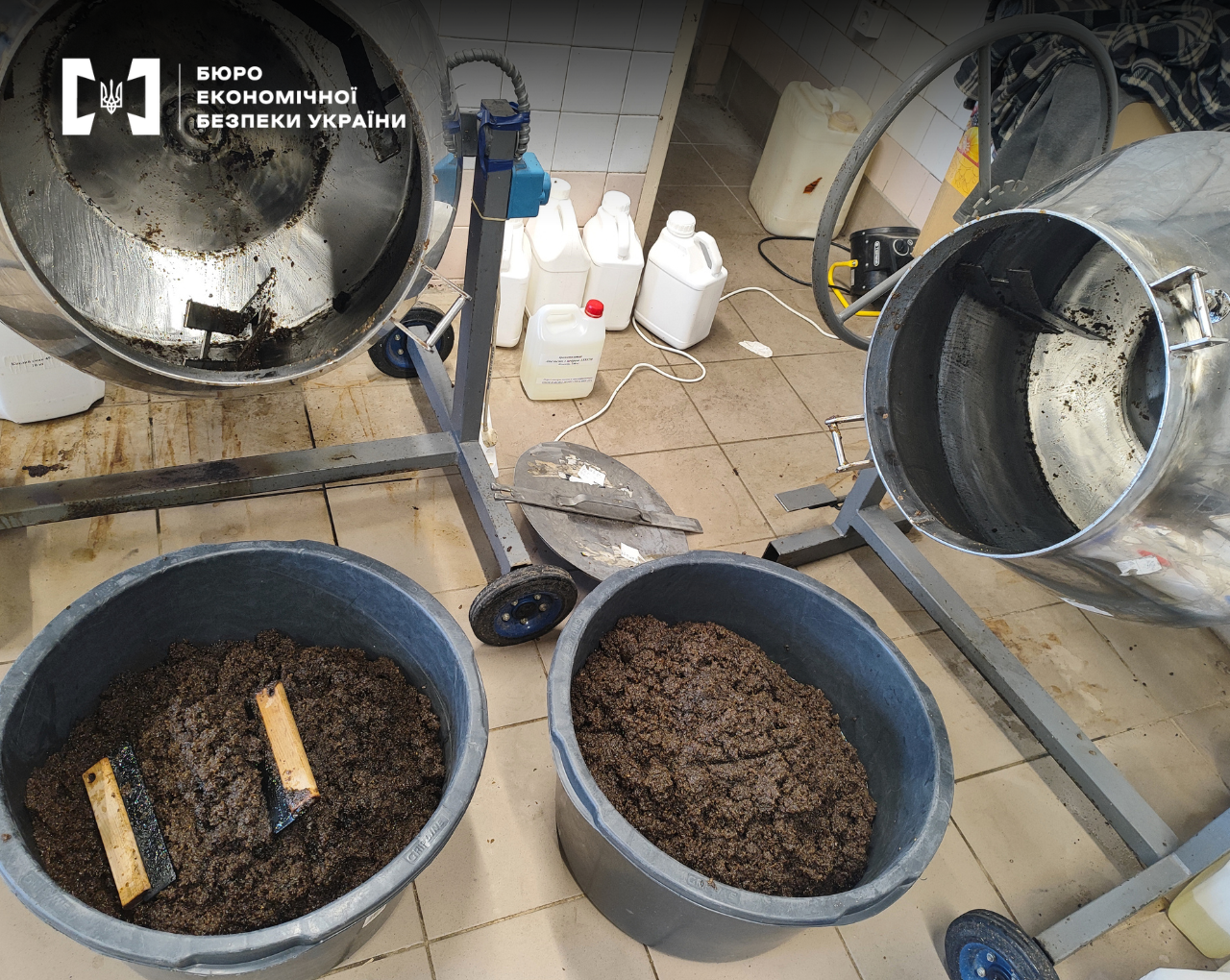 Two large industrial stainless steel mixers, with plastic vats containing a dark brown mass  underneath. White canisters and a technical room are visible in the background.