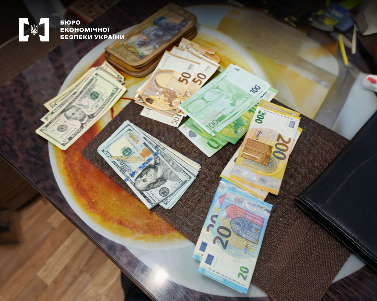 Foreign currency cash laid out on a table. Euro banknotes in denominations of 200, 100, 50, and 20, along with US dollars in denominations of 5 and 100. A leather wallet is next to them