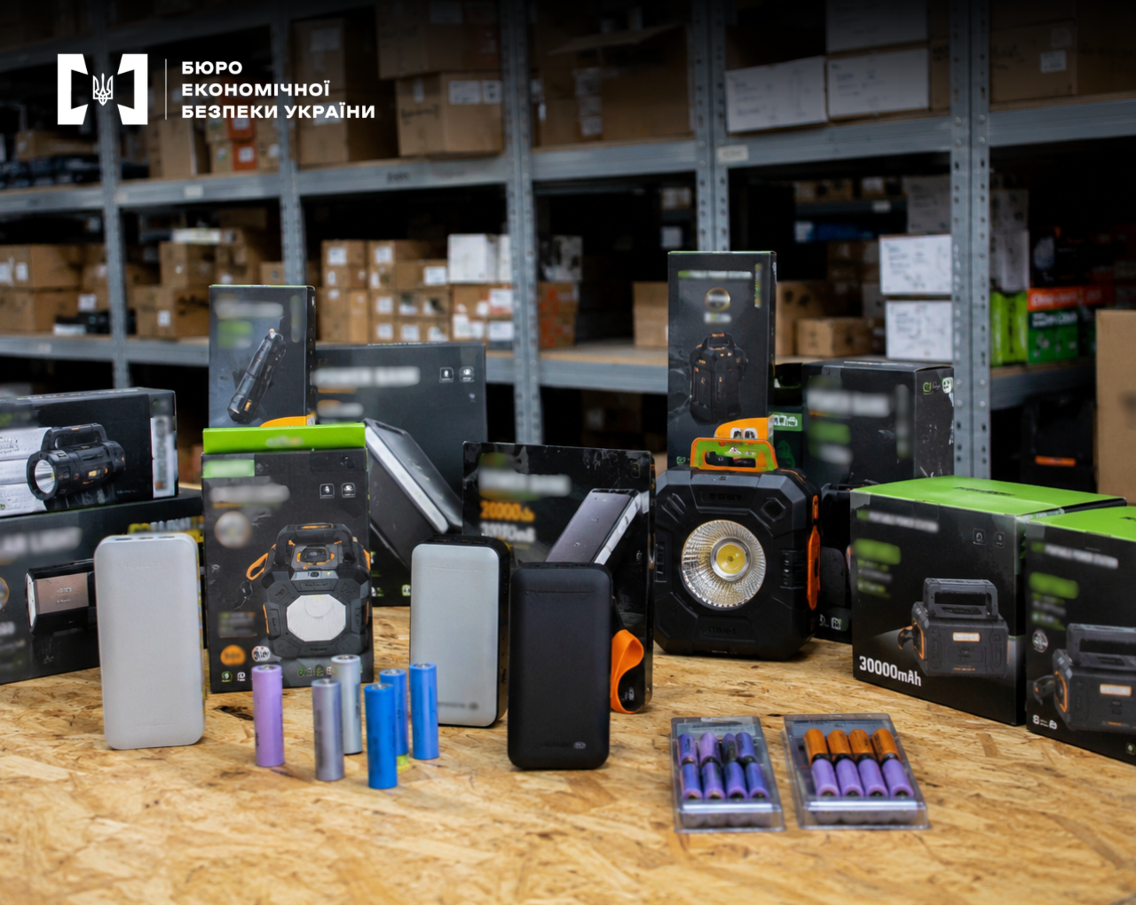 Various portable electronics, power banks, flashlights, and rechargeable batteries are arranged on a wooden table against a background of warehouse shelving units filled with boxes