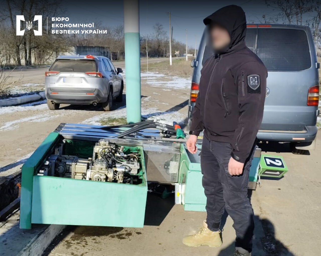 ESBU detective stands next to a dismantled green fuel dispenser. Nearby, there are two vehicles: a grey crossover and a minibus.