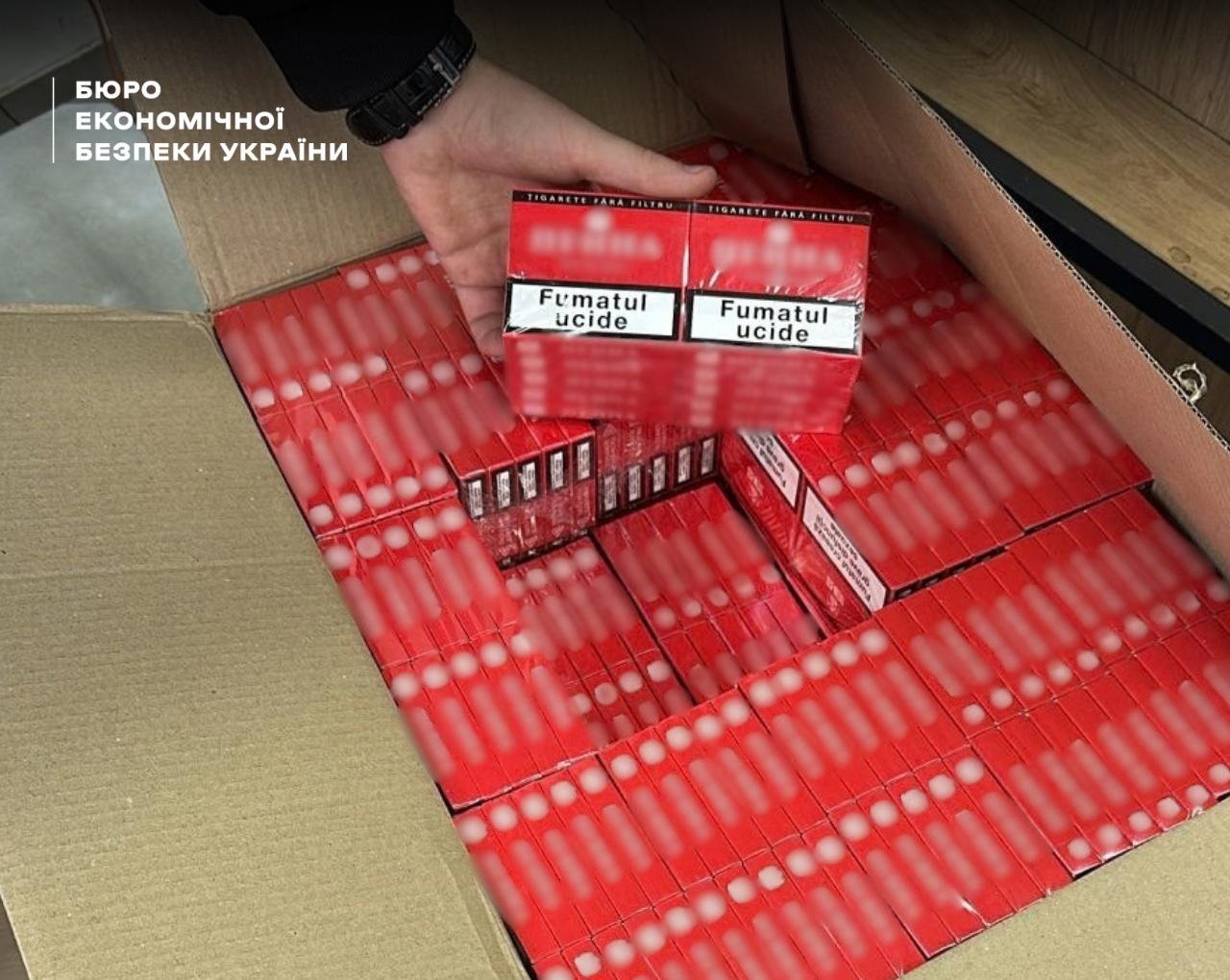A hand in a black jacket holds two red packs of cigarettes with Romanian health warnings over a box full of cartons.