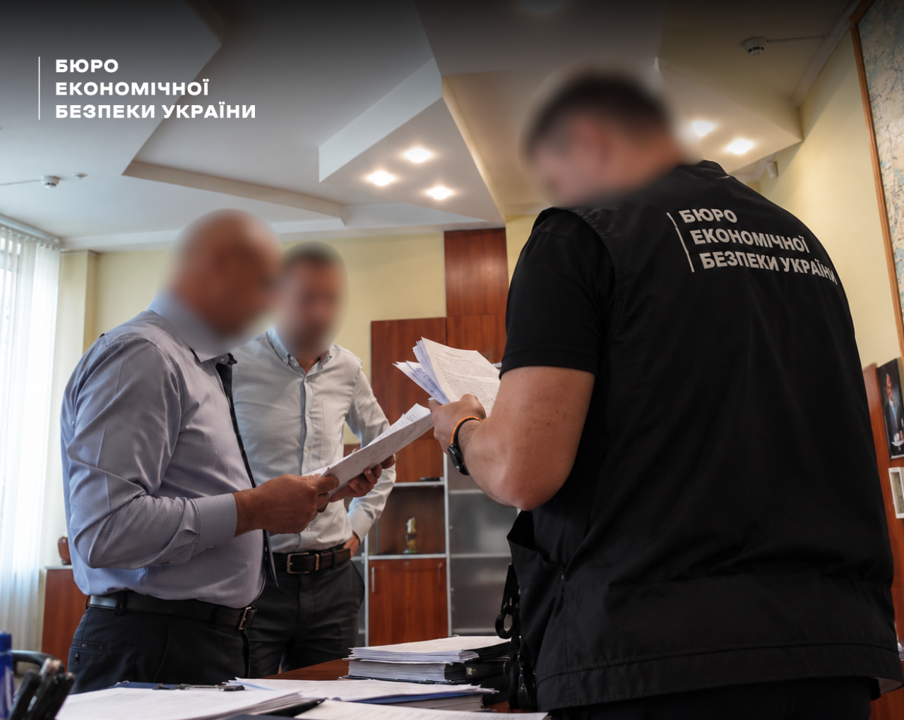 A ESBU detective with their back to the camera holds stacks of papers in front of two men in shirts against the background of a wooden cabinet in an office.