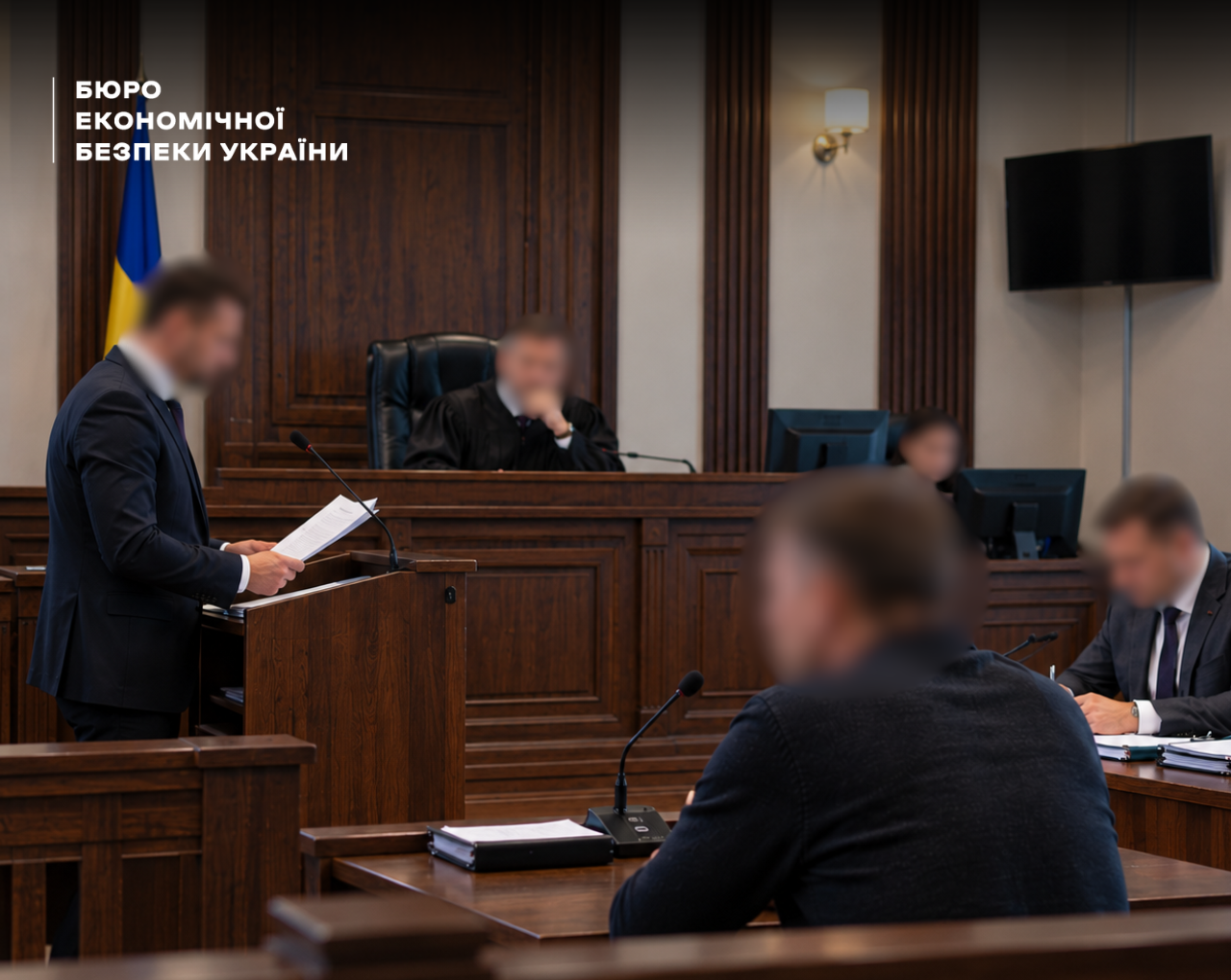 Прокурор за трибуною та суддя на тлі прапора України та дерев'яного оздоблення стін суду