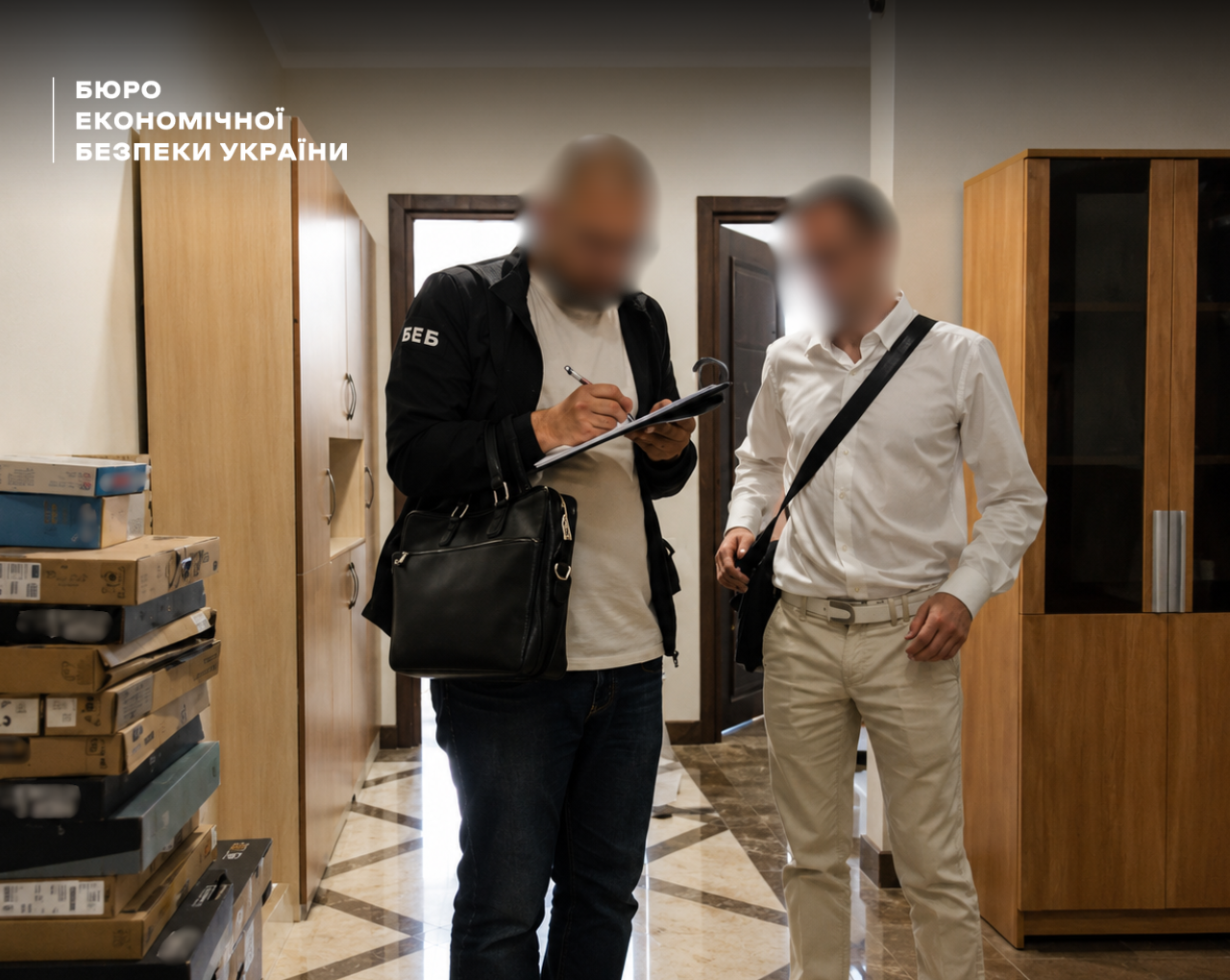 An ESBU detective holding a tablet and a man in a white shirt stand in an office corridor next to stacks of boxes containing branded laptops.