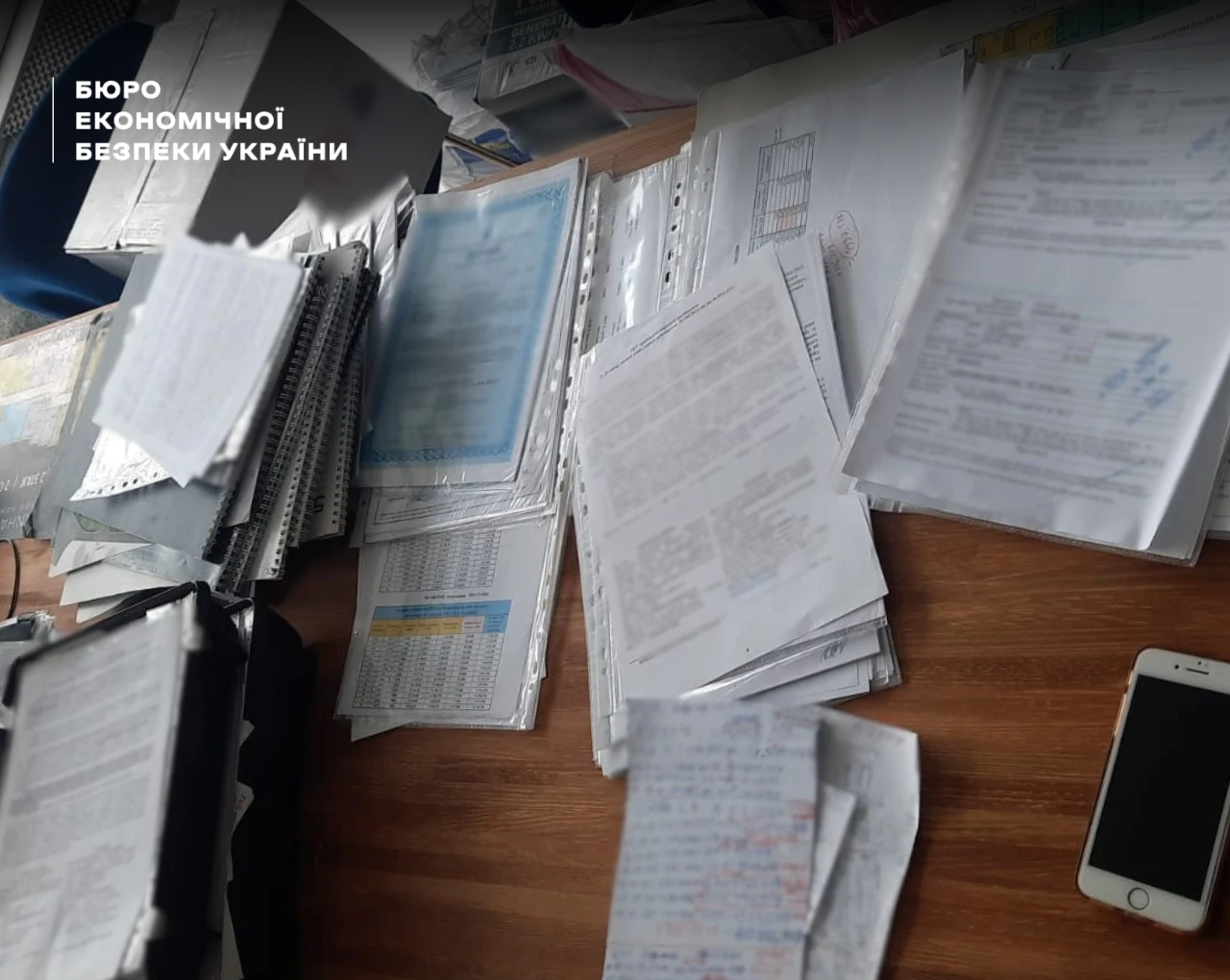 A stack of printed documents, folders, forms, and a mobile phone on a wooden table