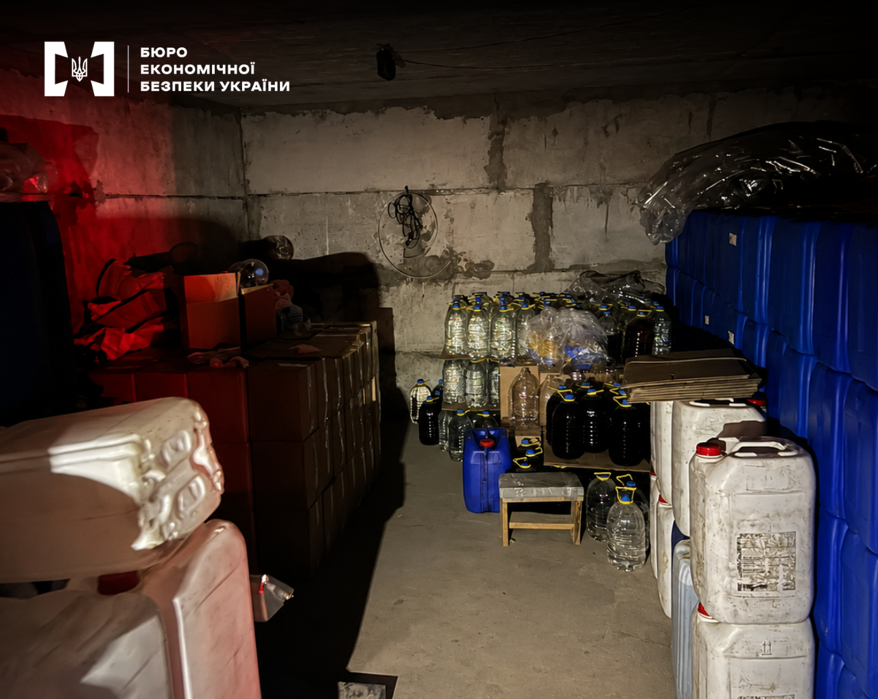 A dark room with concrete walls, filled with canisters, large bottles of liquid, and cardboard boxes.