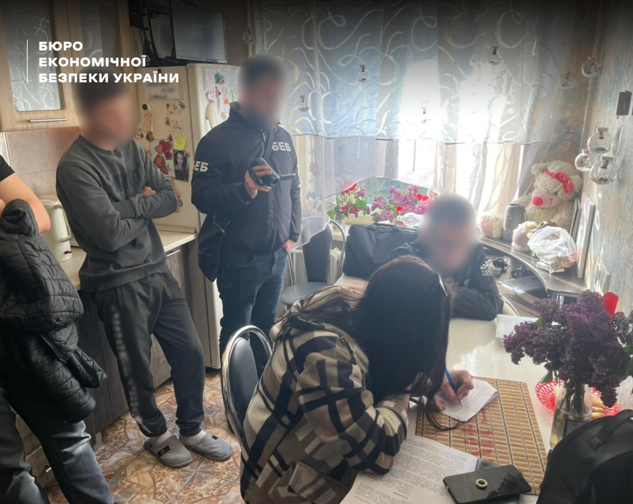 The interior of a kitchen in a residential apartment. A woman wearing a cardigan sits at the table signing documents, next to men whose faces are blurred. Behind them stands an ESBU detective holding a device.