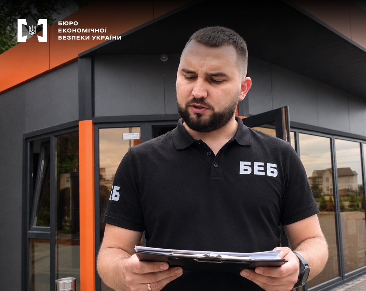 A close-up of an ESBU detective holding a clipboard with papers. In the background is a modern, single-story gas station building with dark tinted glass