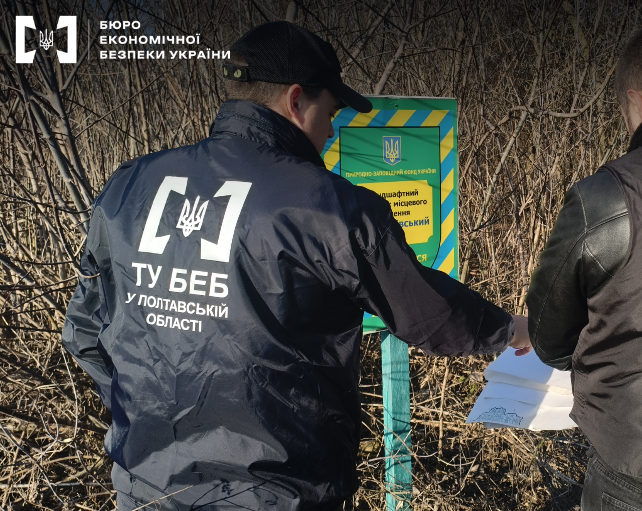 An open area. An ESBU detective hands documents to a man in the background near a boundary marker of a nature reserve fund.