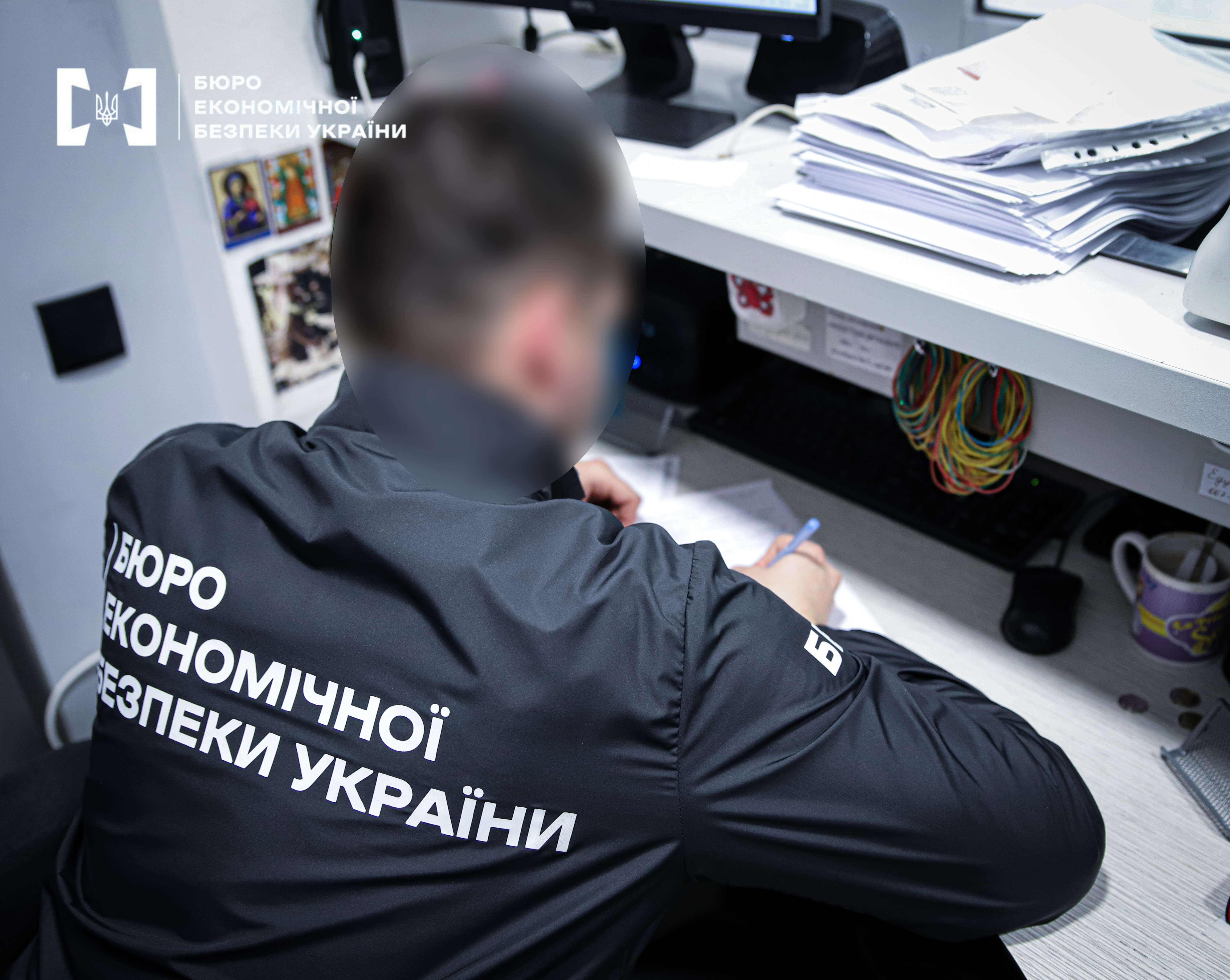 An ESBU detective is sitting at a desk. Their face is blurred. The detective is working with documents, with a large stack of papers and a computer monitor nearby.