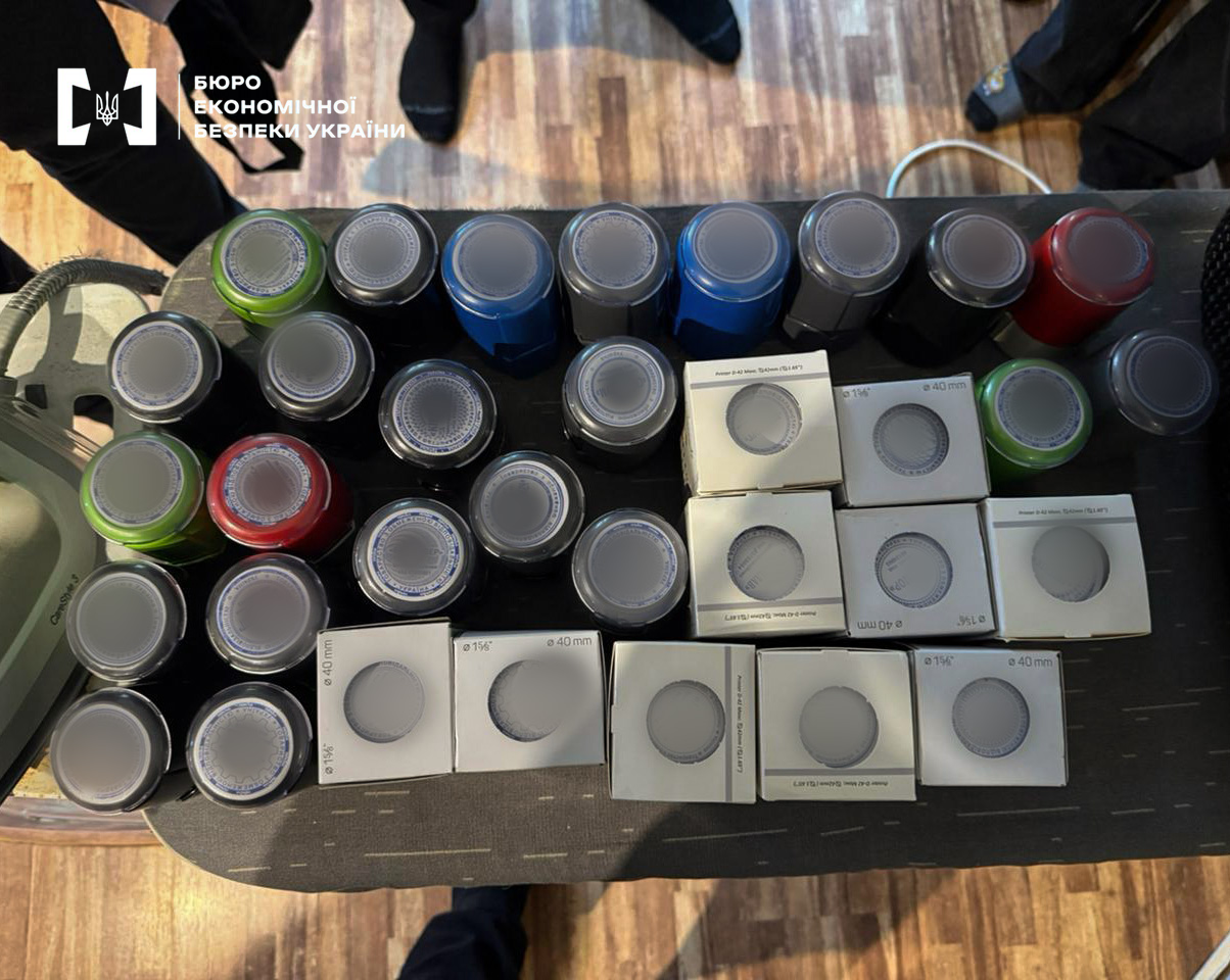 A top view of a large number of official seals and stamps in various colors (blue, red, black, and green) spread out on a table. Cardboard boxes for stamp mounts are lying nearby.