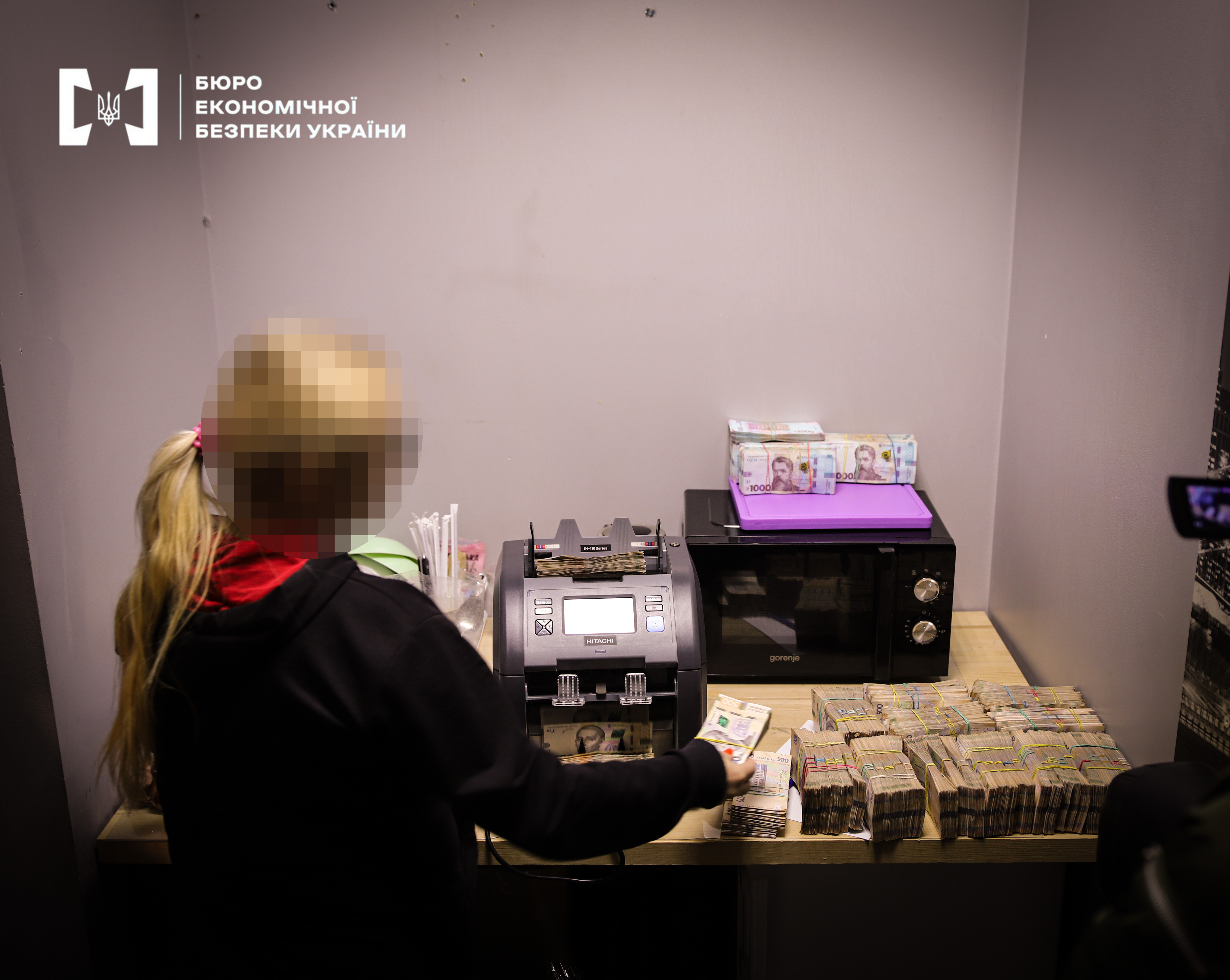 A woman stands before a desk equipped with a money-counting machine. She is holding a banknote in her hands. Numerous stacks of cash are lying on the desk and on top of a microwave oven
