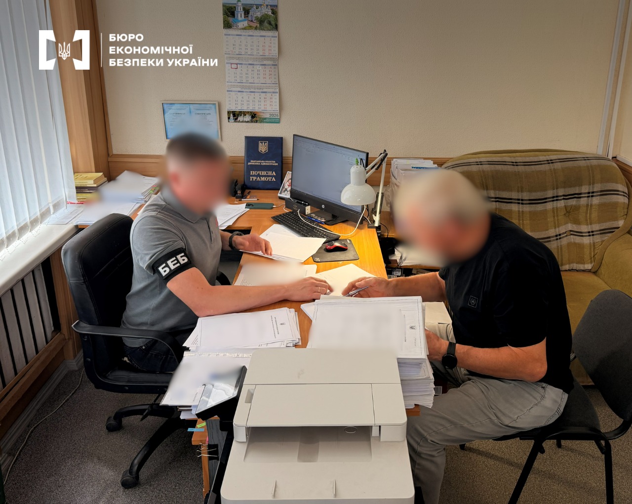 A ESBU detective briefs a suspect in the case on the investigation materials in an office near a printer. The faces of the people are blurred.