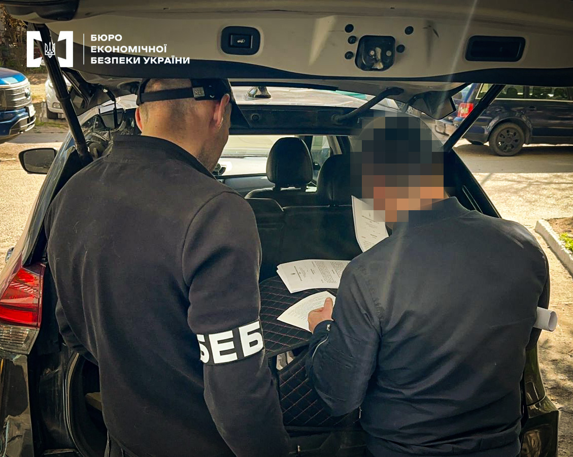 ESBU detectives are conducting investigative actions. They are standing near the rear of a car with the tailgate open, holding documents in their hands.