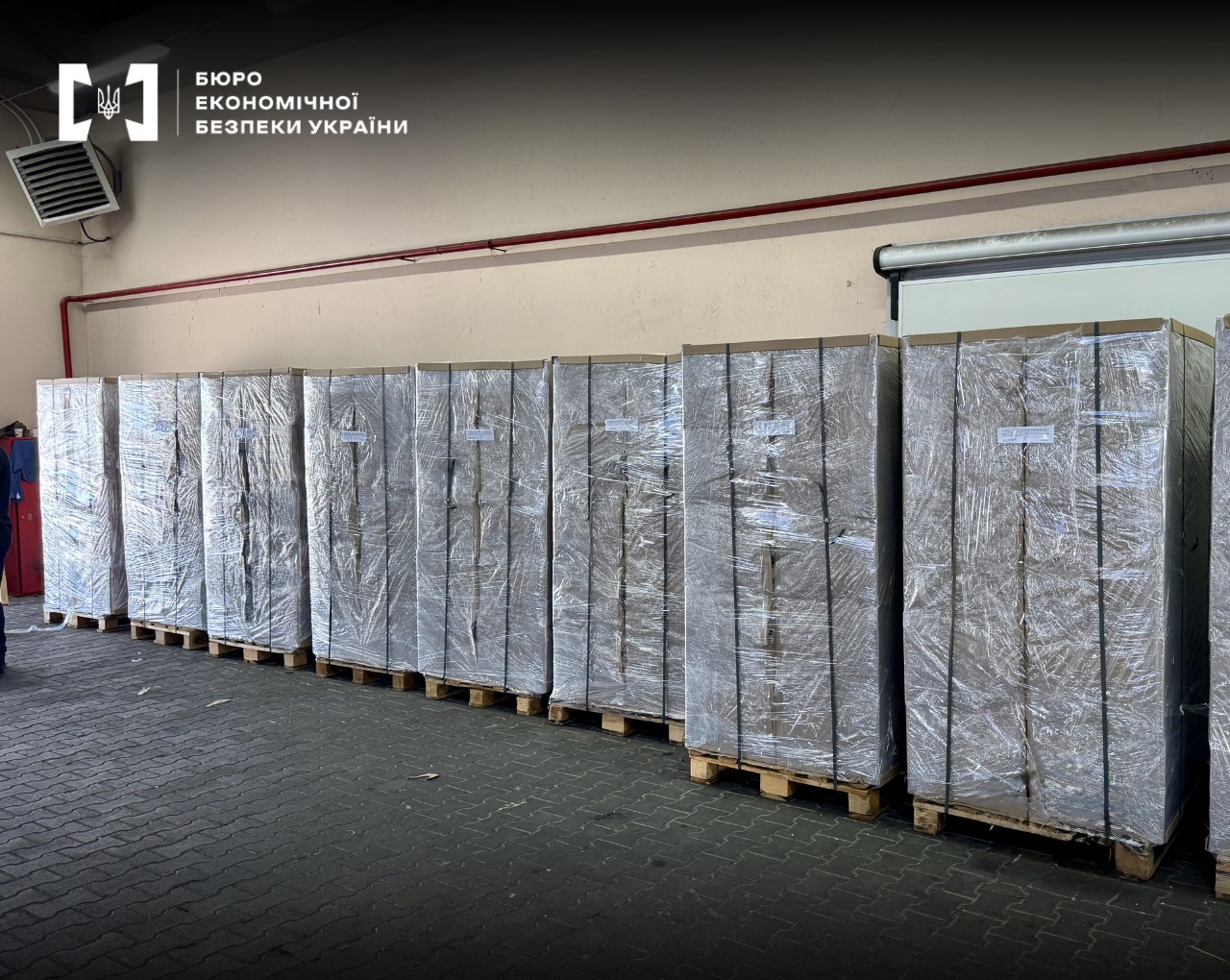A row of large pallets with cargo wrapped in transparent film along a wall in a warehouse