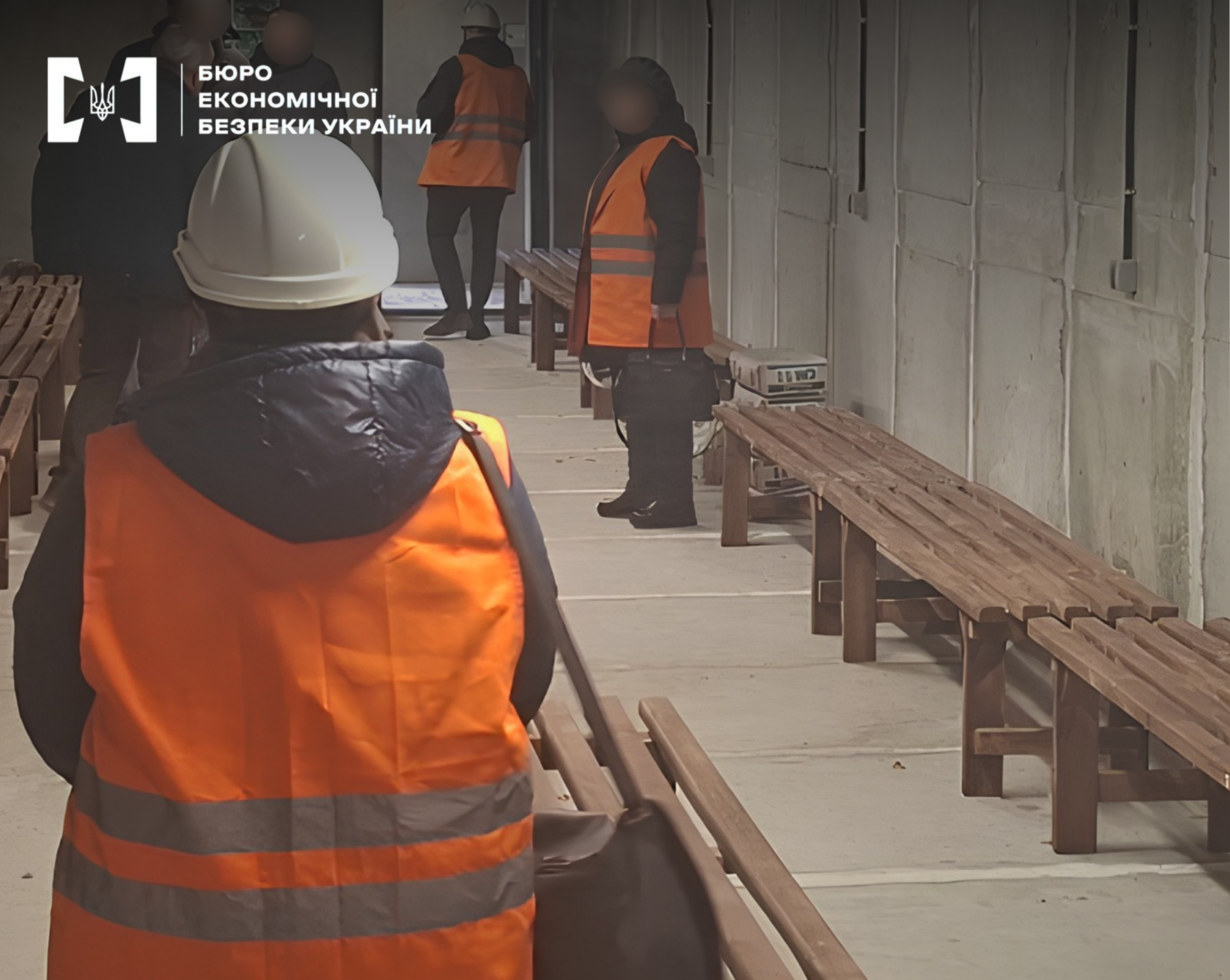People in construction hard hats and orange vests inspect an underground shelter with wooden benches along the walls.