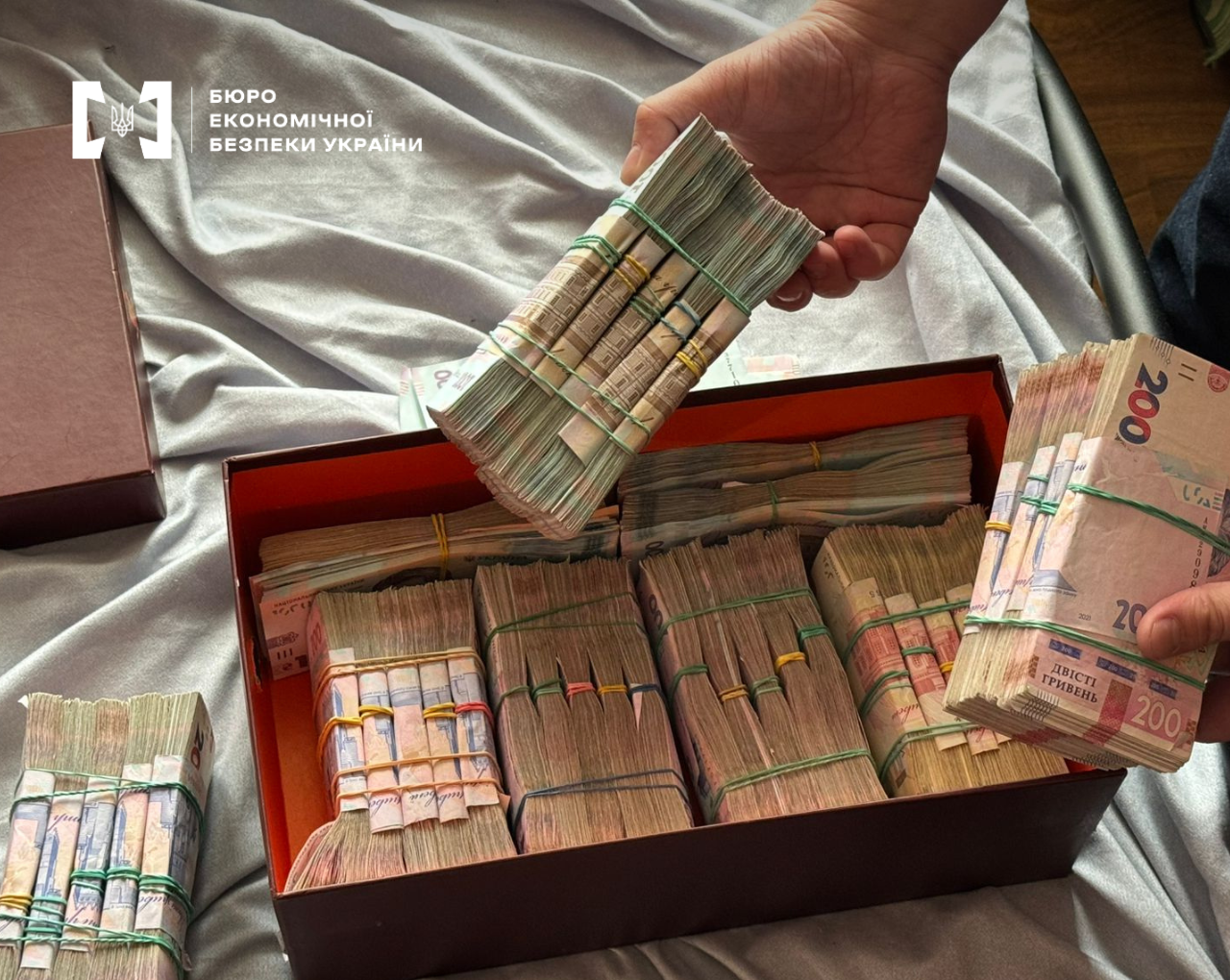 A large cardboard box filled with thick stacks of Ukrainian hryvnia cash. Two people are holding additional bundles of 200 and 500 denomination banknotes