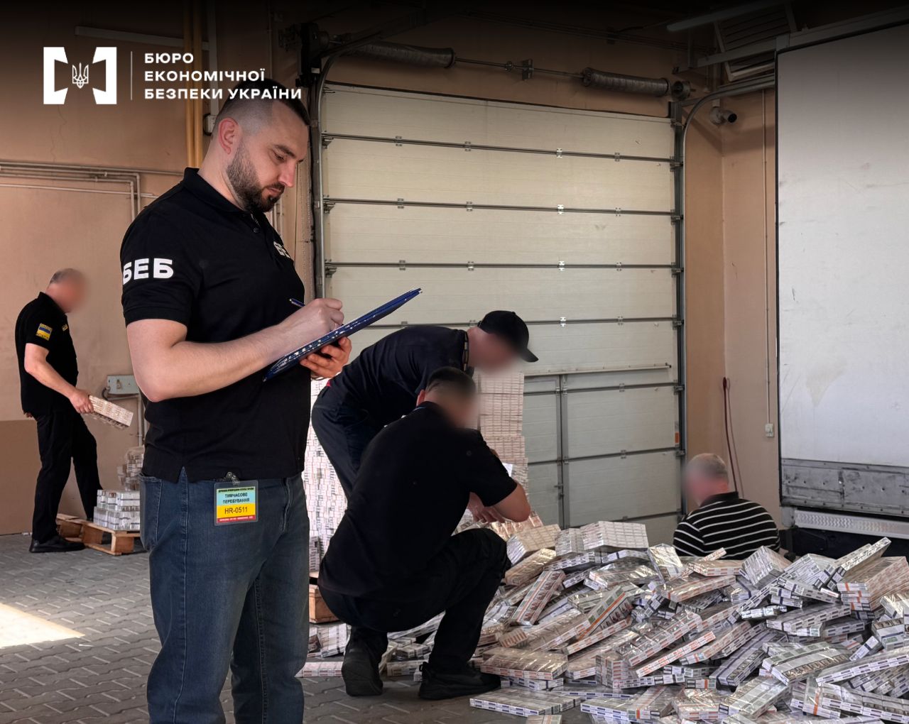 A ESBU detective in a black polo shirt takes notes on a tablet while other employees count a huge pile of seized packs of smuggled cigarettes.
