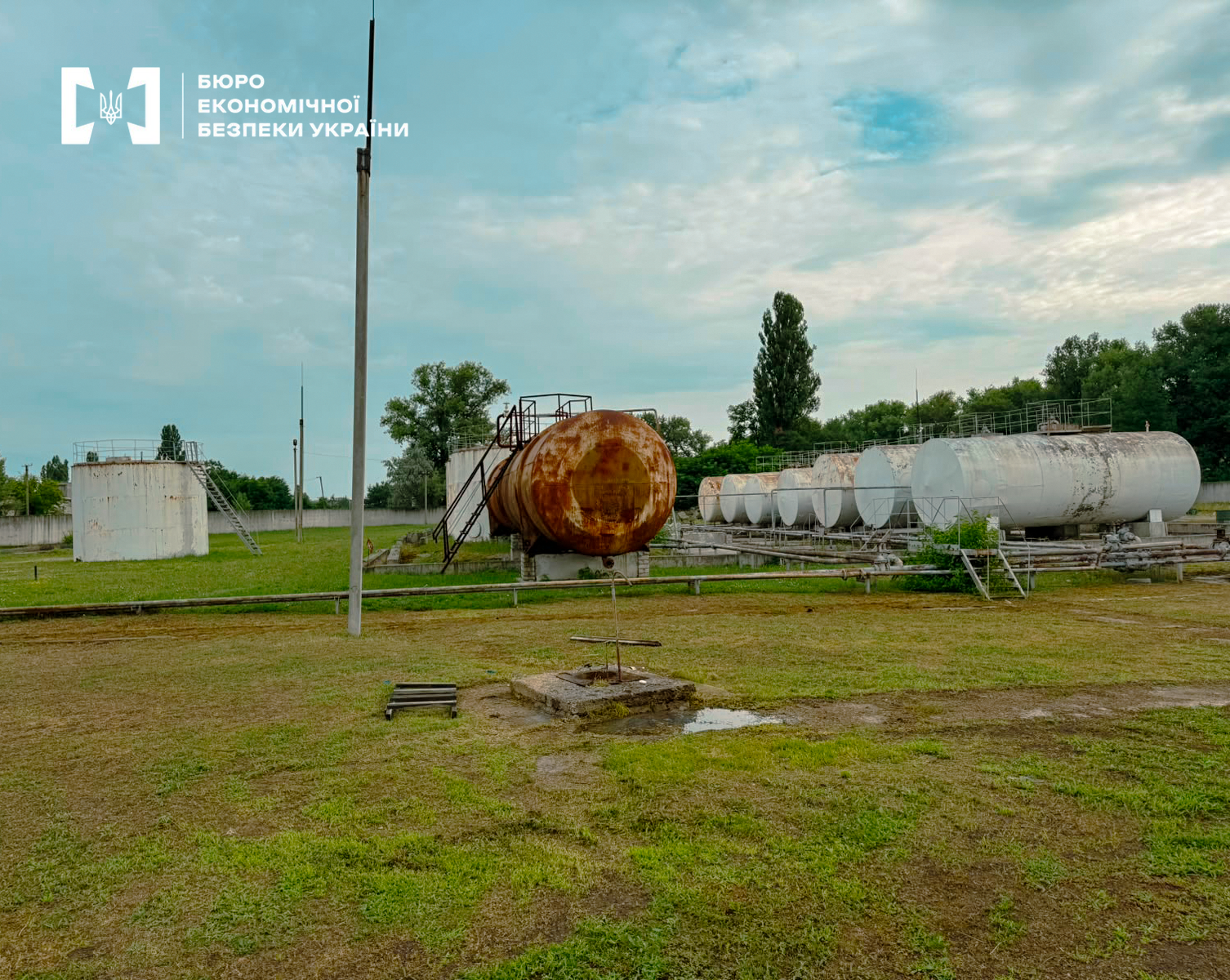 A row of large horizontal tanks and a rusty reservoir on the territory of a base.