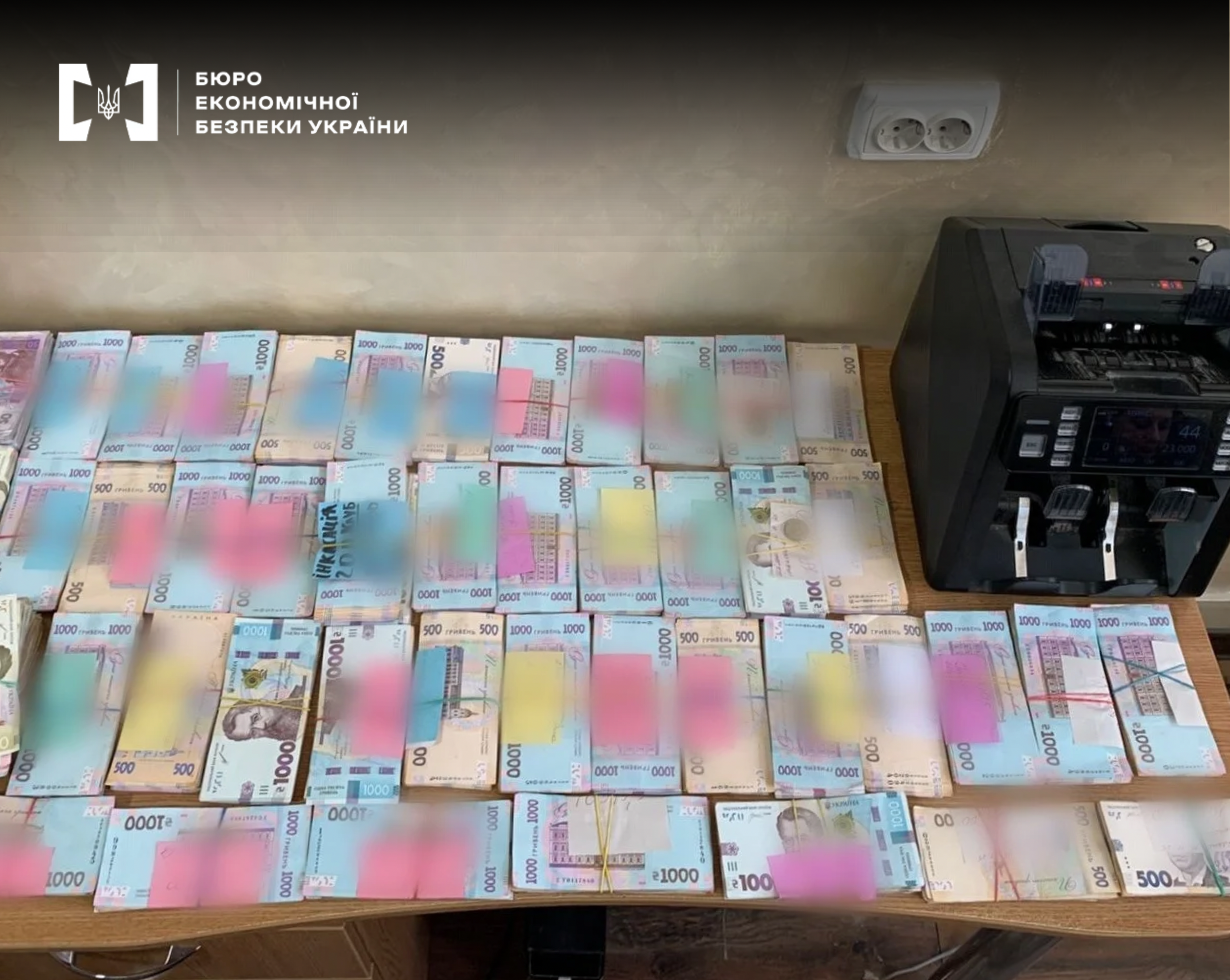 A huge number of bundles of hryvnias of various denominations, arranged in rows next to a banknote counter.
