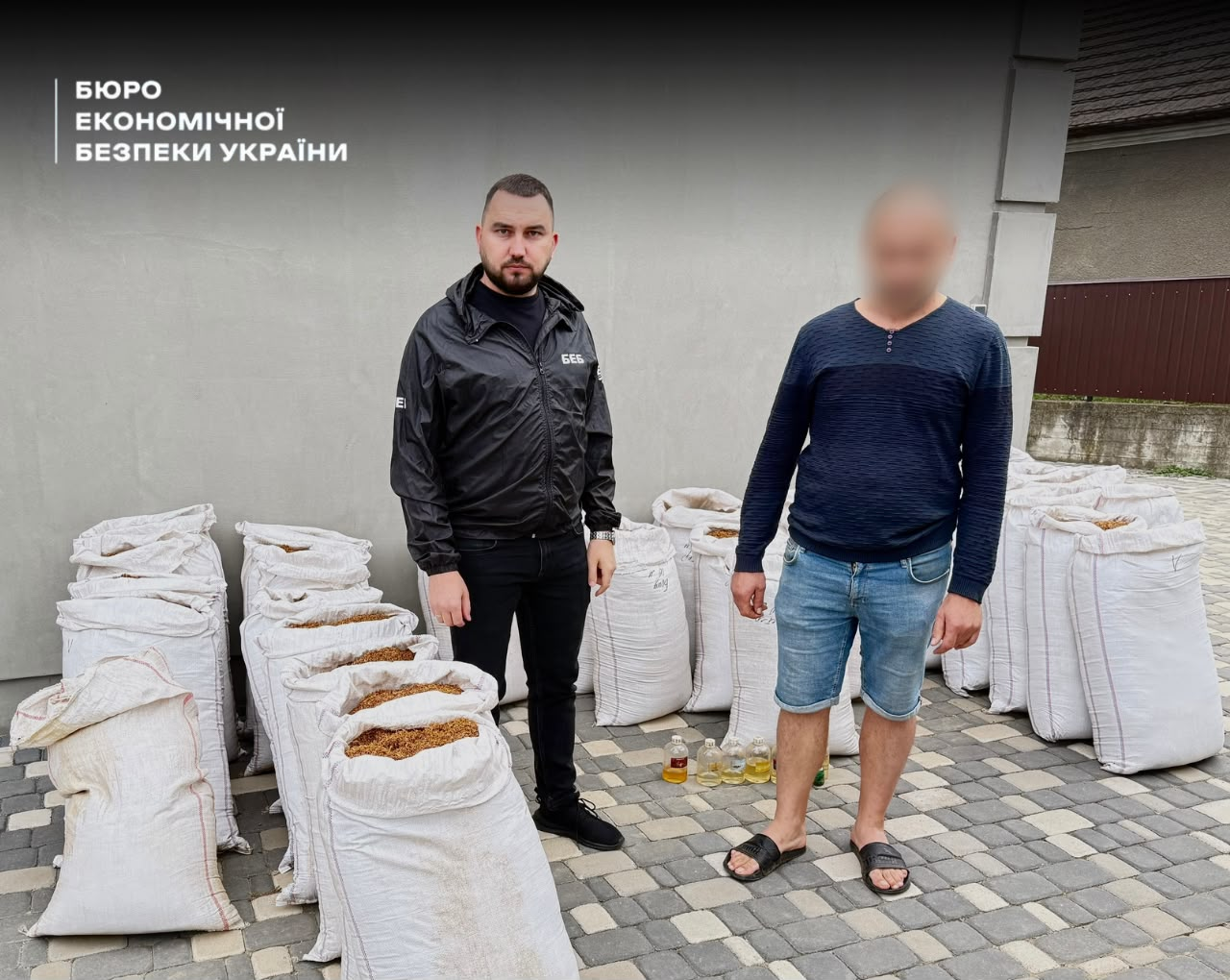 A ESBU detective and the suspect stand in a yard next to dozens of bags of tobacco. The faces are blurred.