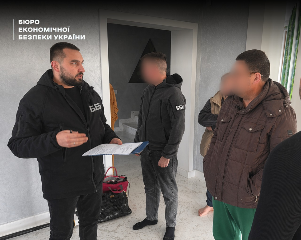 A ESBU detective with documents, an SSU operative, and suspects in the hallway of a private house during the inventory and seizure of property.