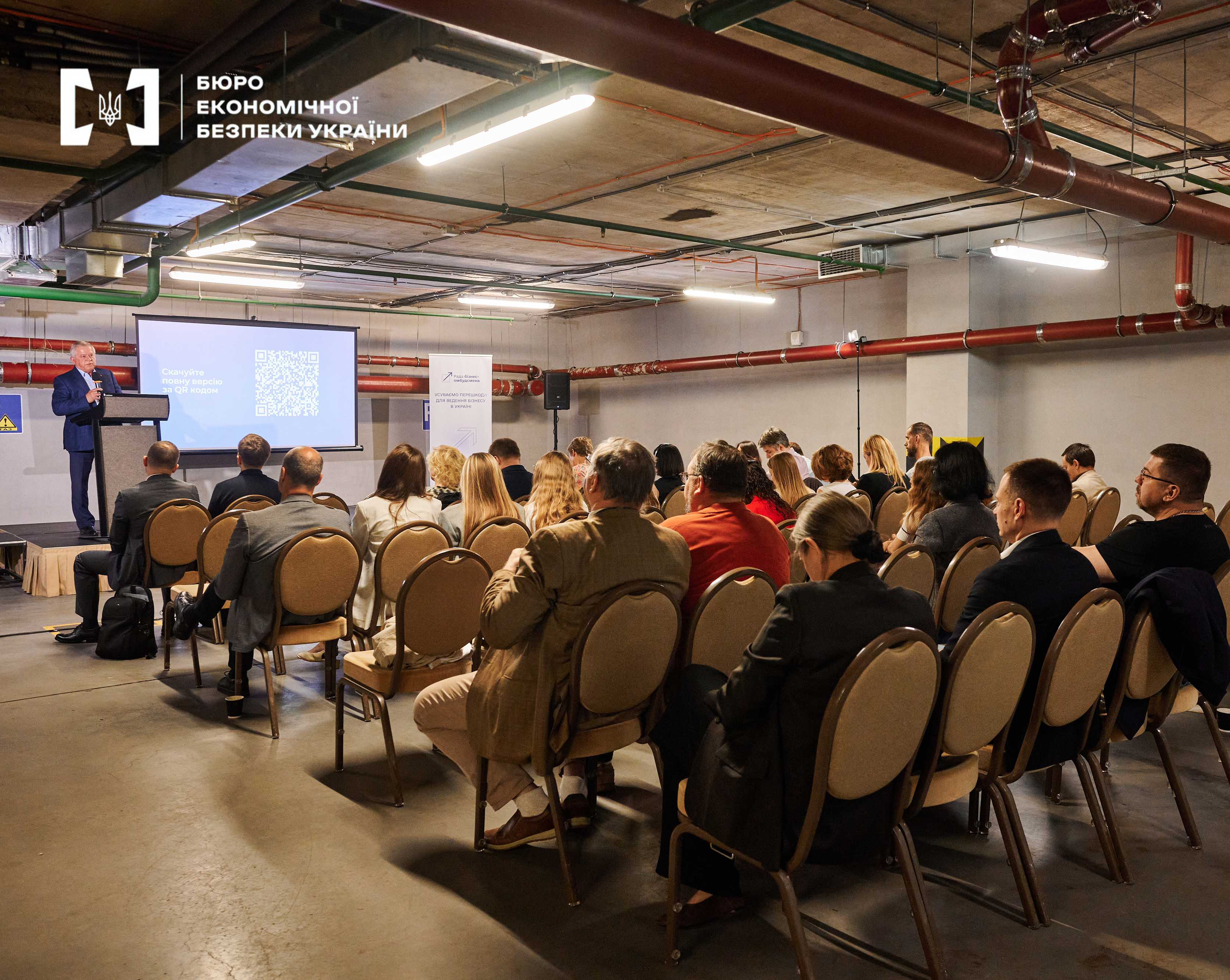 Rows of listeners in a hall with exposed utility pipes during a report.