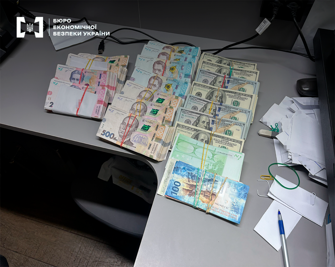 Stacks of banknotes of 200, 500 hryvnias, and 100 US dollars next to a ballpoint pen and forms during the recording of evidence.
