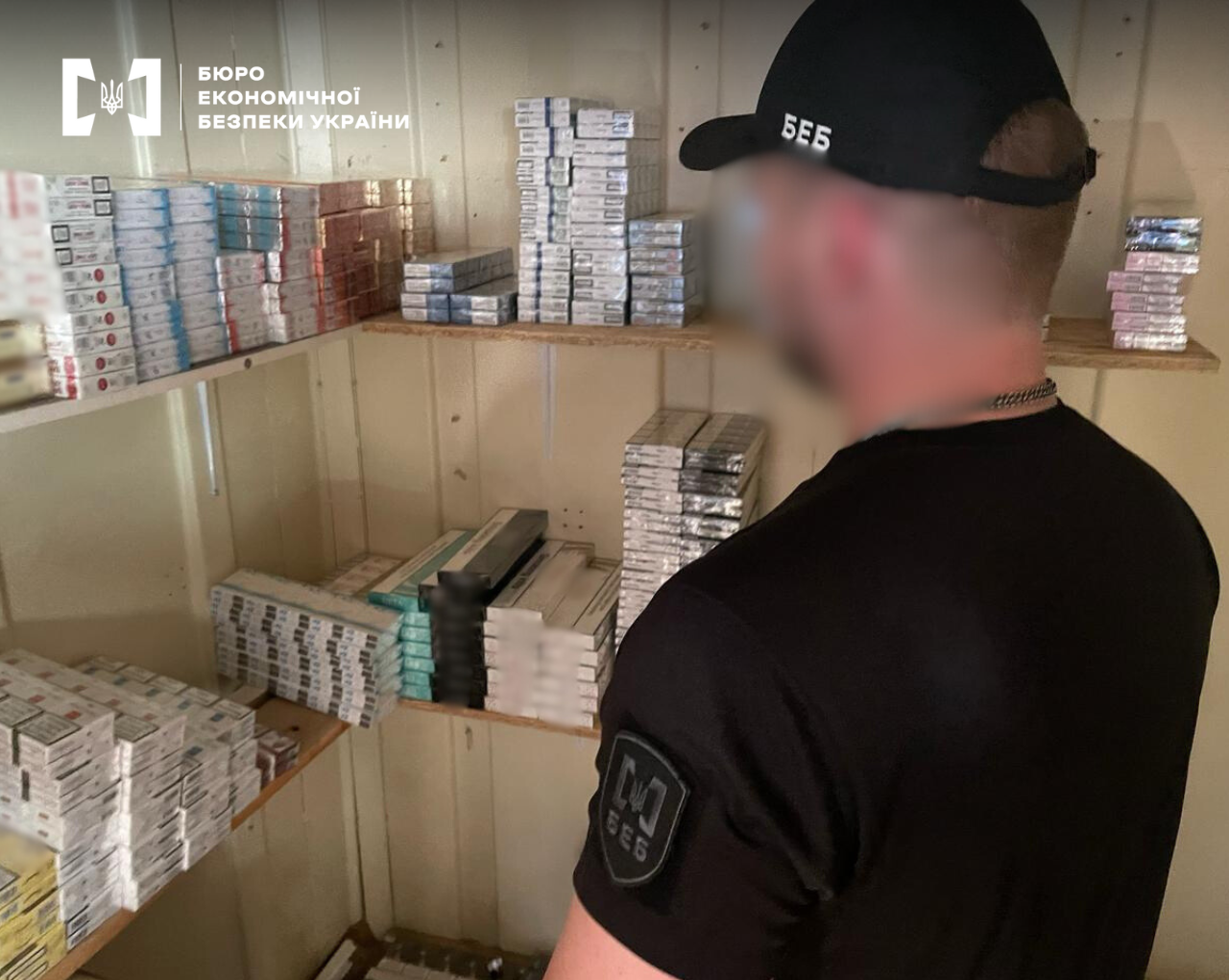 A ESBU detective with their back to the camera examines counterfeit packs of cigarettes on wooden shelves in an underground warehouse.