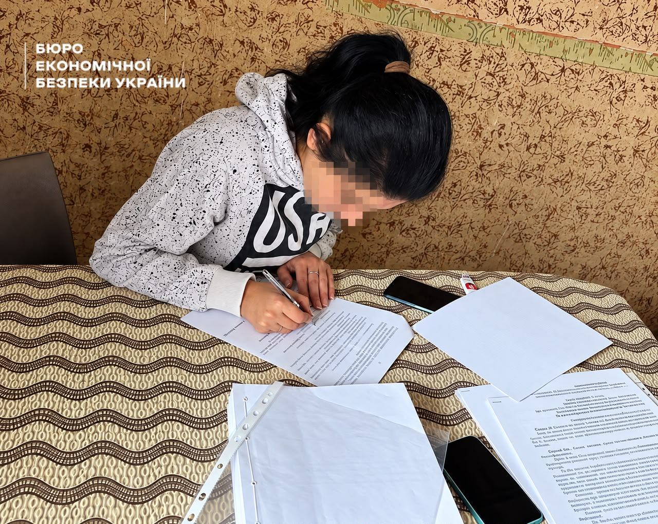 A young woman in a grey sweatshirt fills out papers at a desk where ESBU procedural acts are laid out alongside smartphones. 
