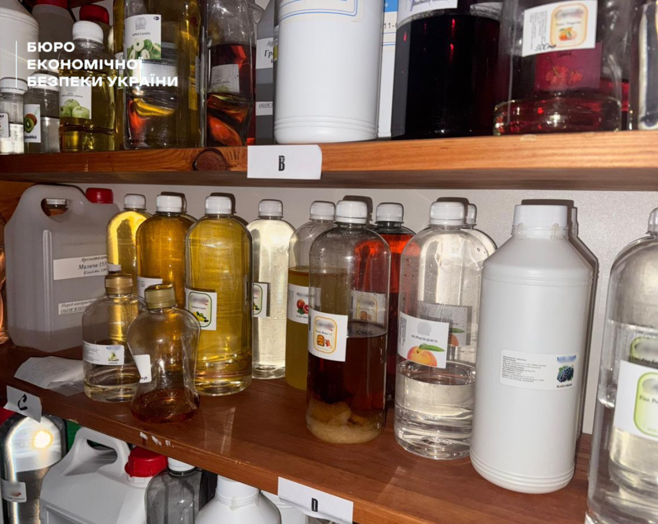 Plastic bottles with multi-coloured essences (peach, grape, apple) on wooden shelving units under indexes 