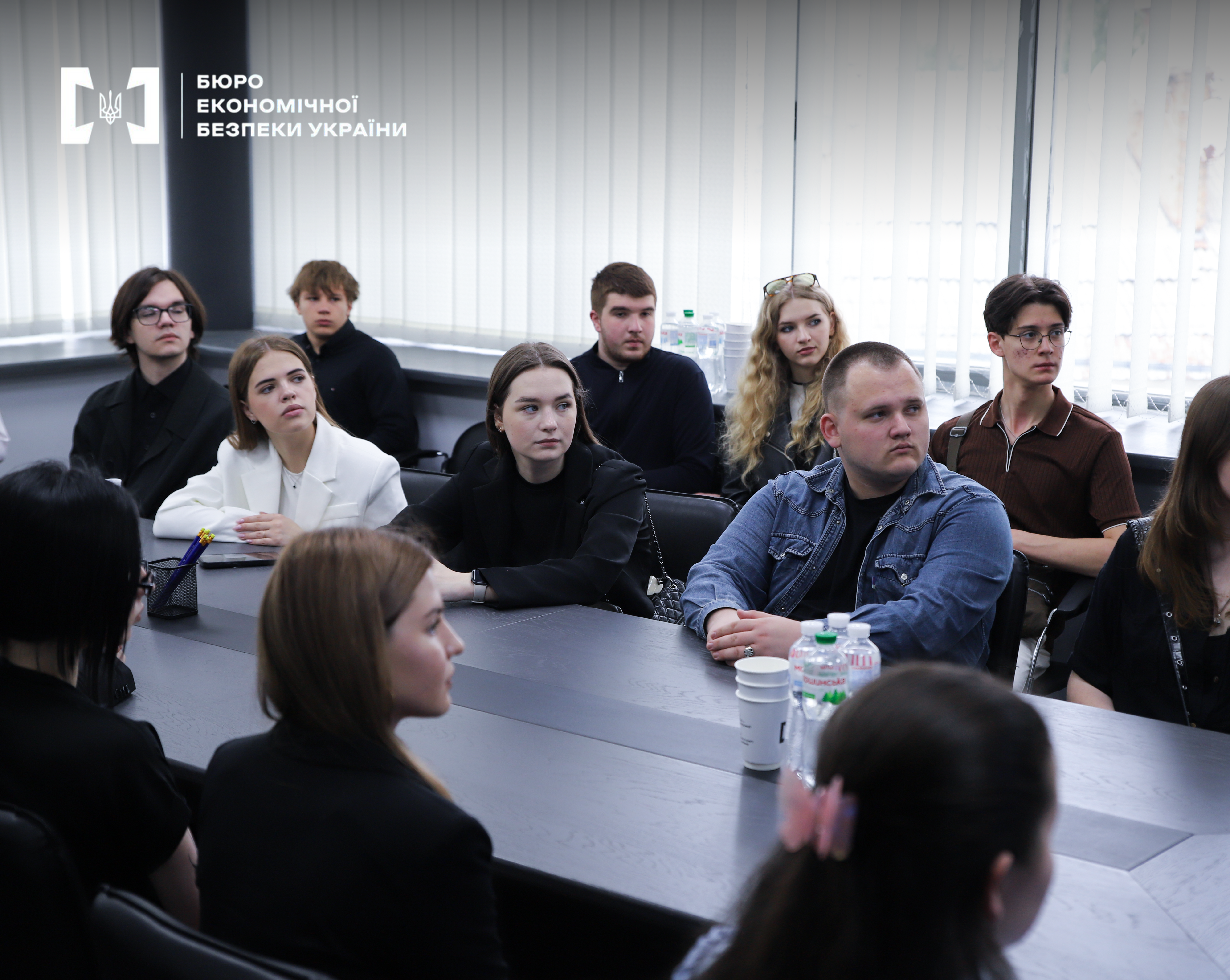 A meeting between the ESBU leadership and youth in a conference hall. The shot features a group of young people at a table, listening attentively to a speaker off-camera