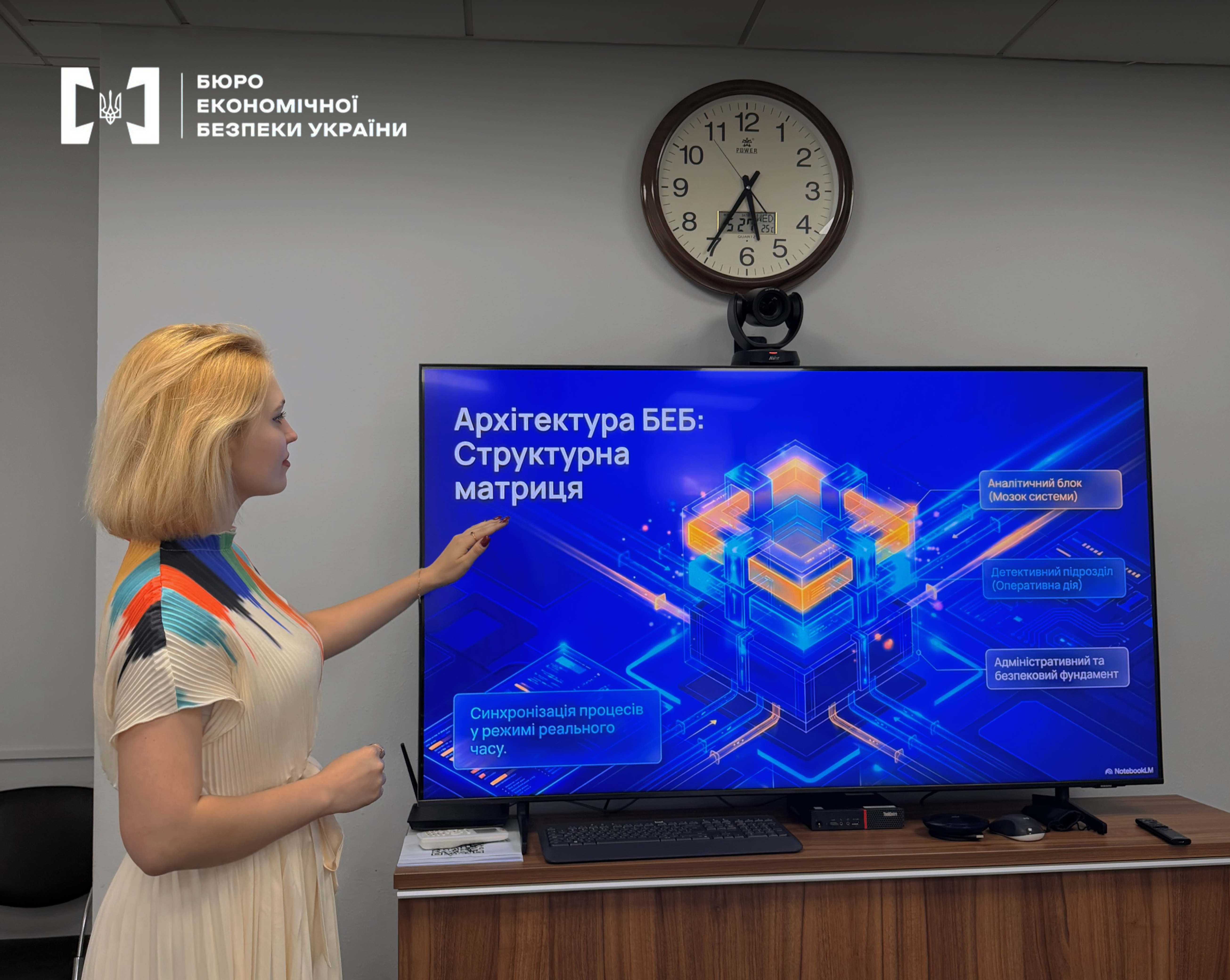 A female speaker demonstrates a presentation titled 'ESBU Architecture: Structural Matrix' on a large screen. A round wall clock is visible above the screen