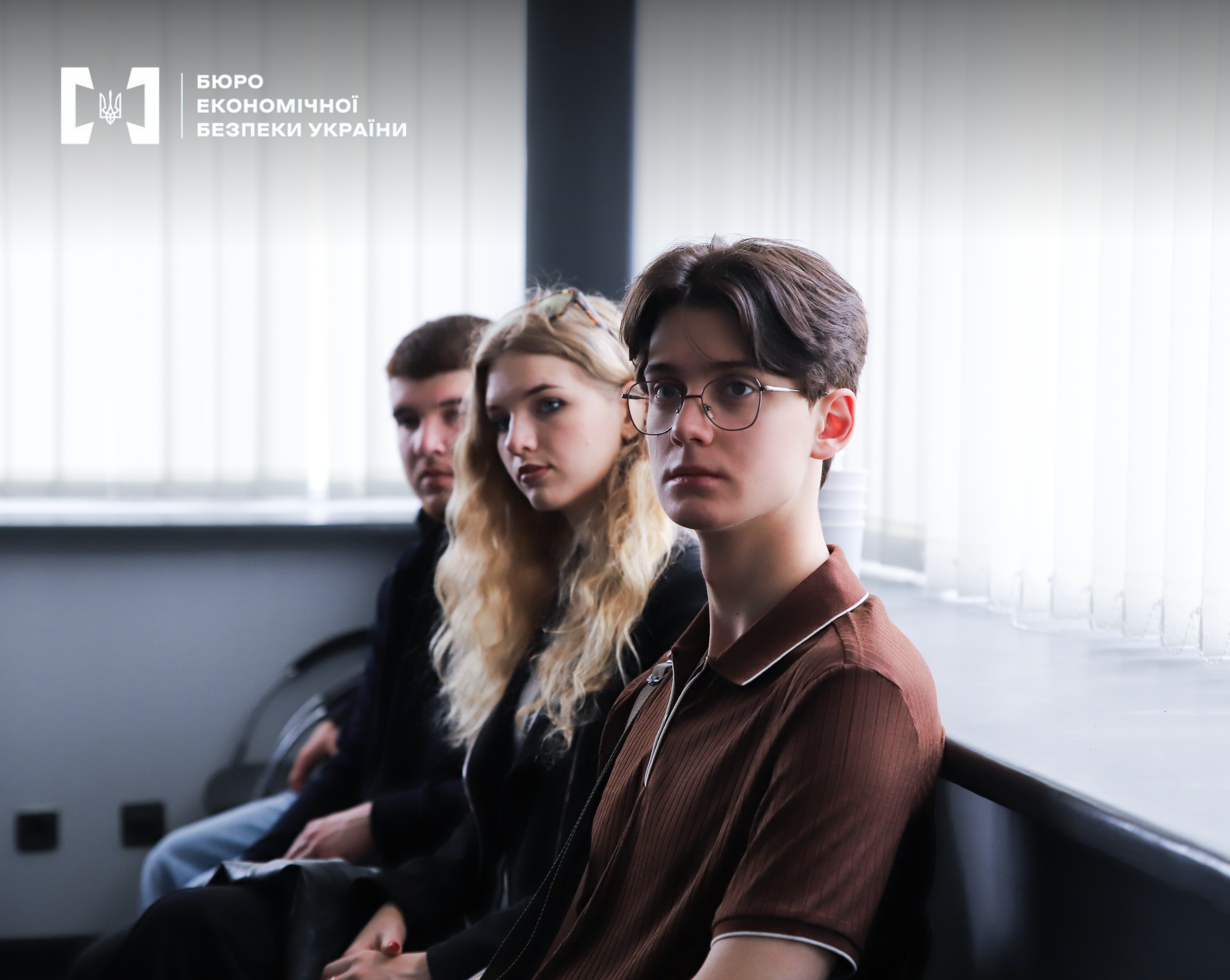 Students listen attentively to a speech at an official meeting in a hall with window blinds. Three people are in the frame—two young men and a young woman with curly blonde hair.